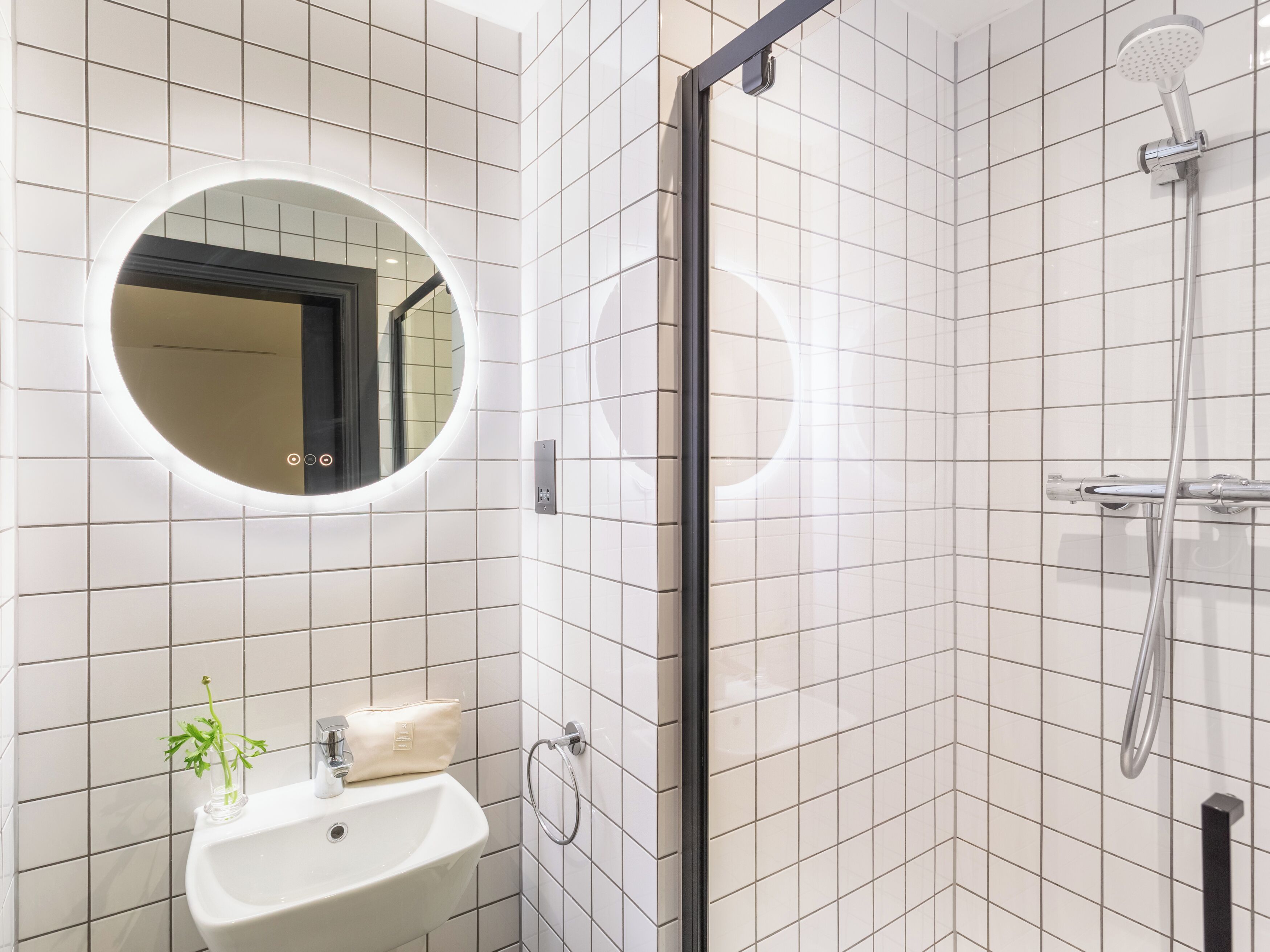 A modern bathroom with white tiled walls, a round mirror, a sink, and a shower.