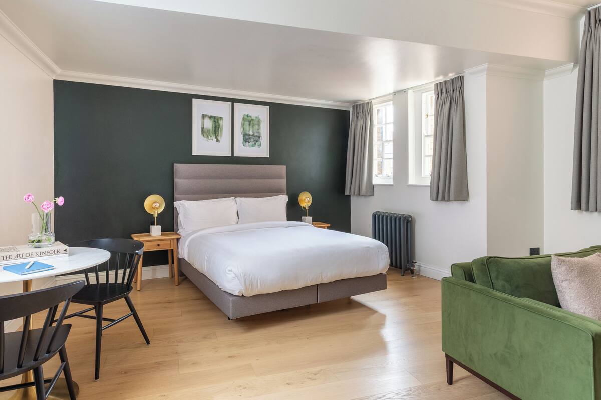 Een slaapkamer met een bed, een groene fauteuil, een klein tafeltje met een stoel en twee bedlampjes.
