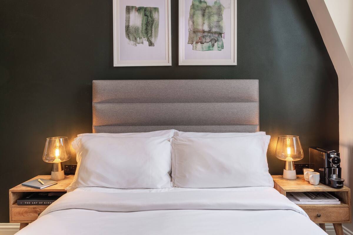 Een net opgemaakt bed met witte lakens, twee bedlampen en een nachtkastje met een boek en een koffiezetapparaat.