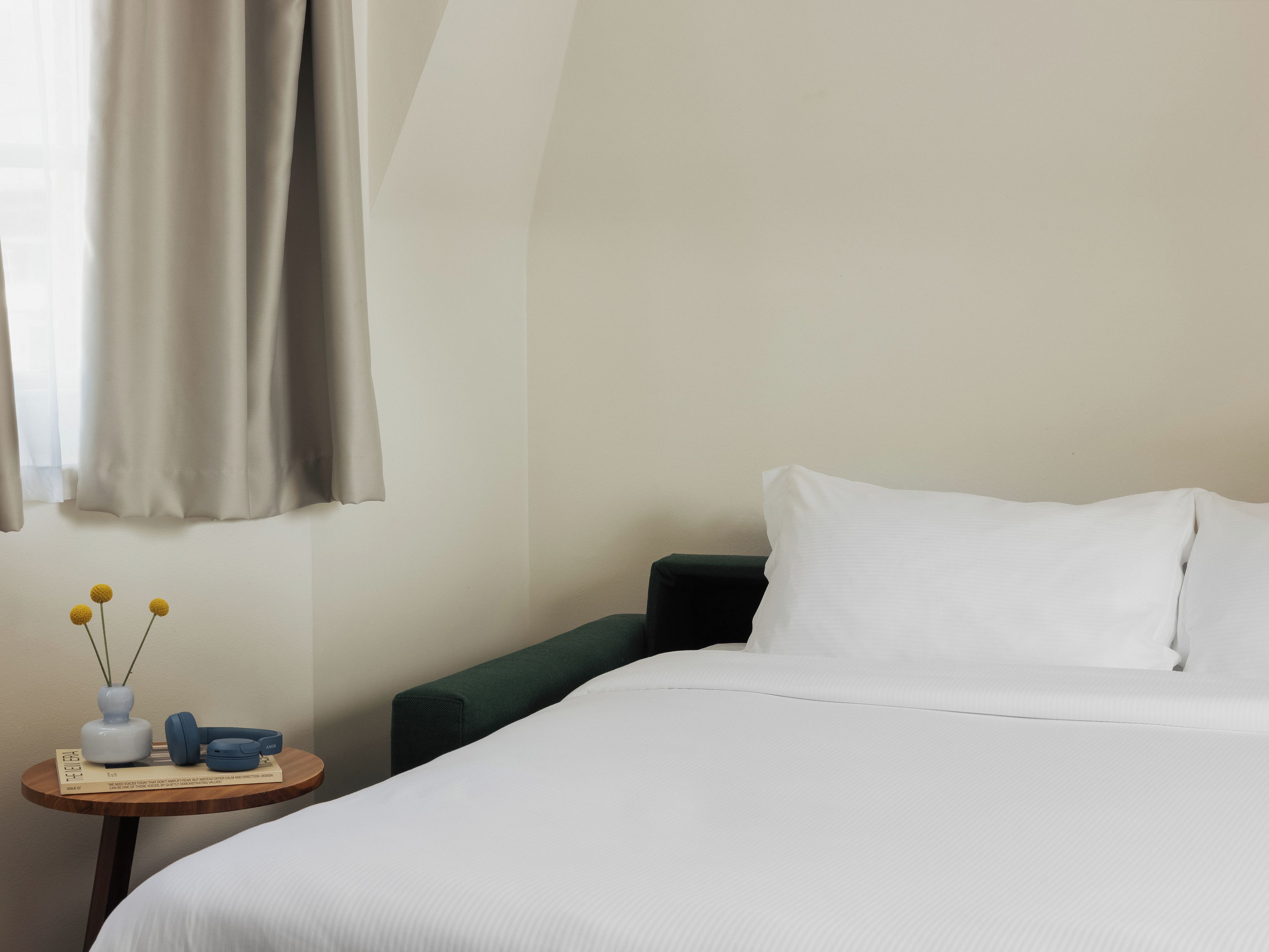 A hotel room with a bed, a small table with a vase and headphones, and a green chair.