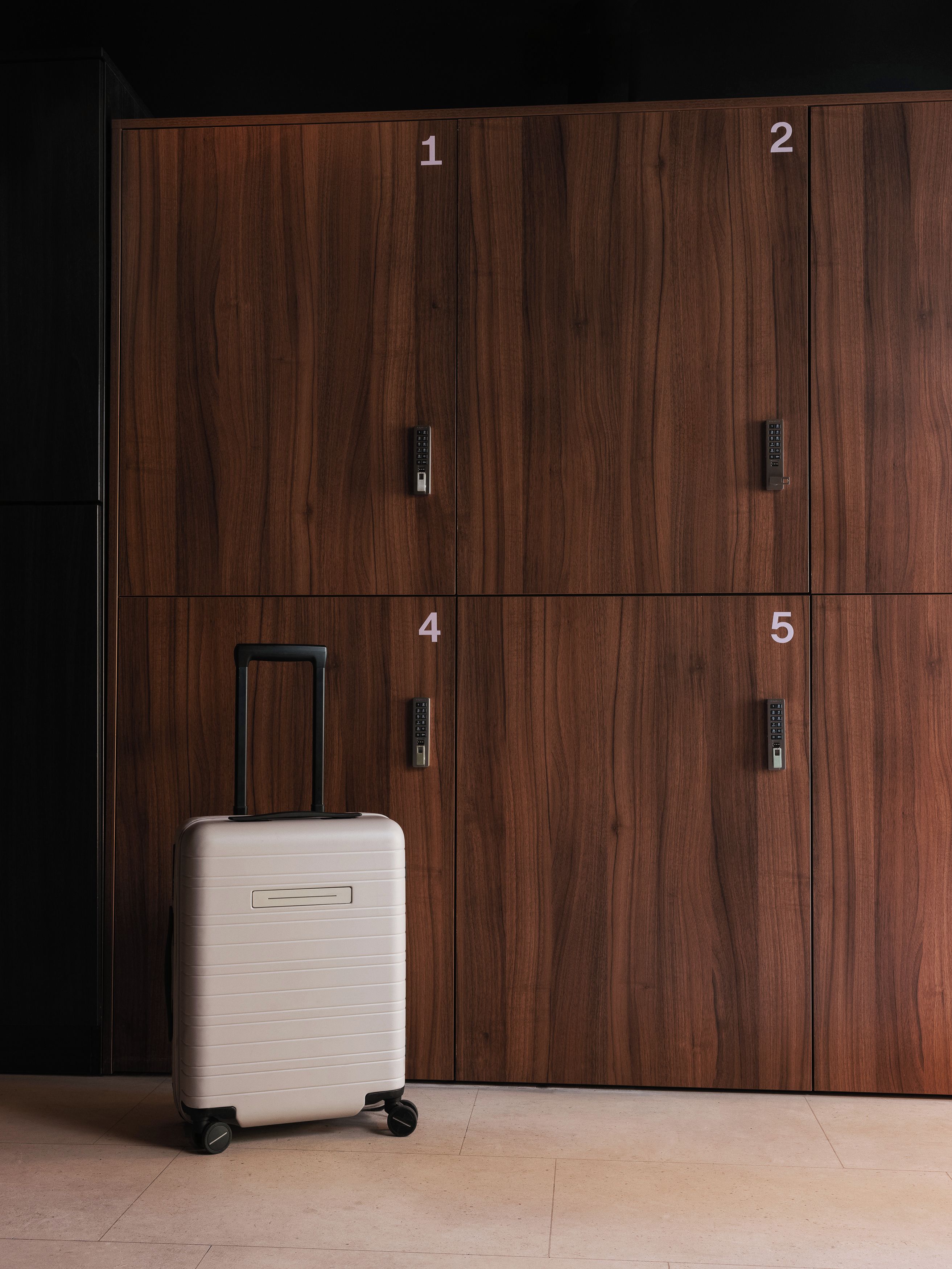 A set of wooden lockers with numbers 1 to 5, a suitcase with wheels, and a remote control on locker number 1.