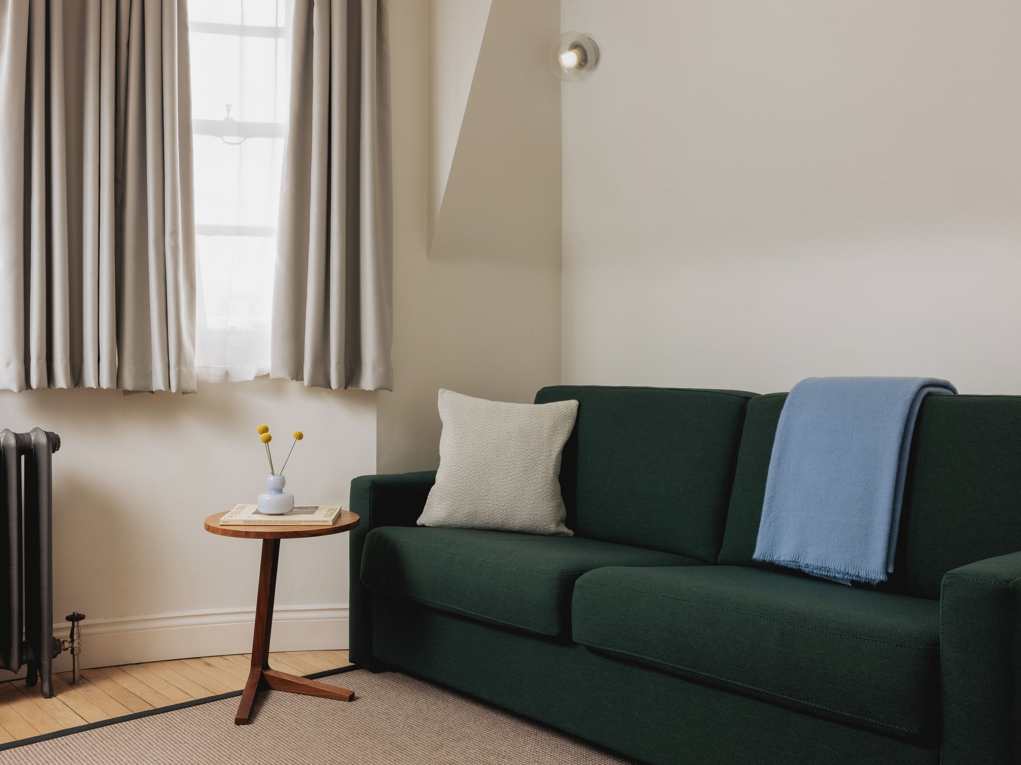 A green sofa with a white pillow and a blue throw blanket, a small wooden side table with a vase and a book, and a radiator on the floor.