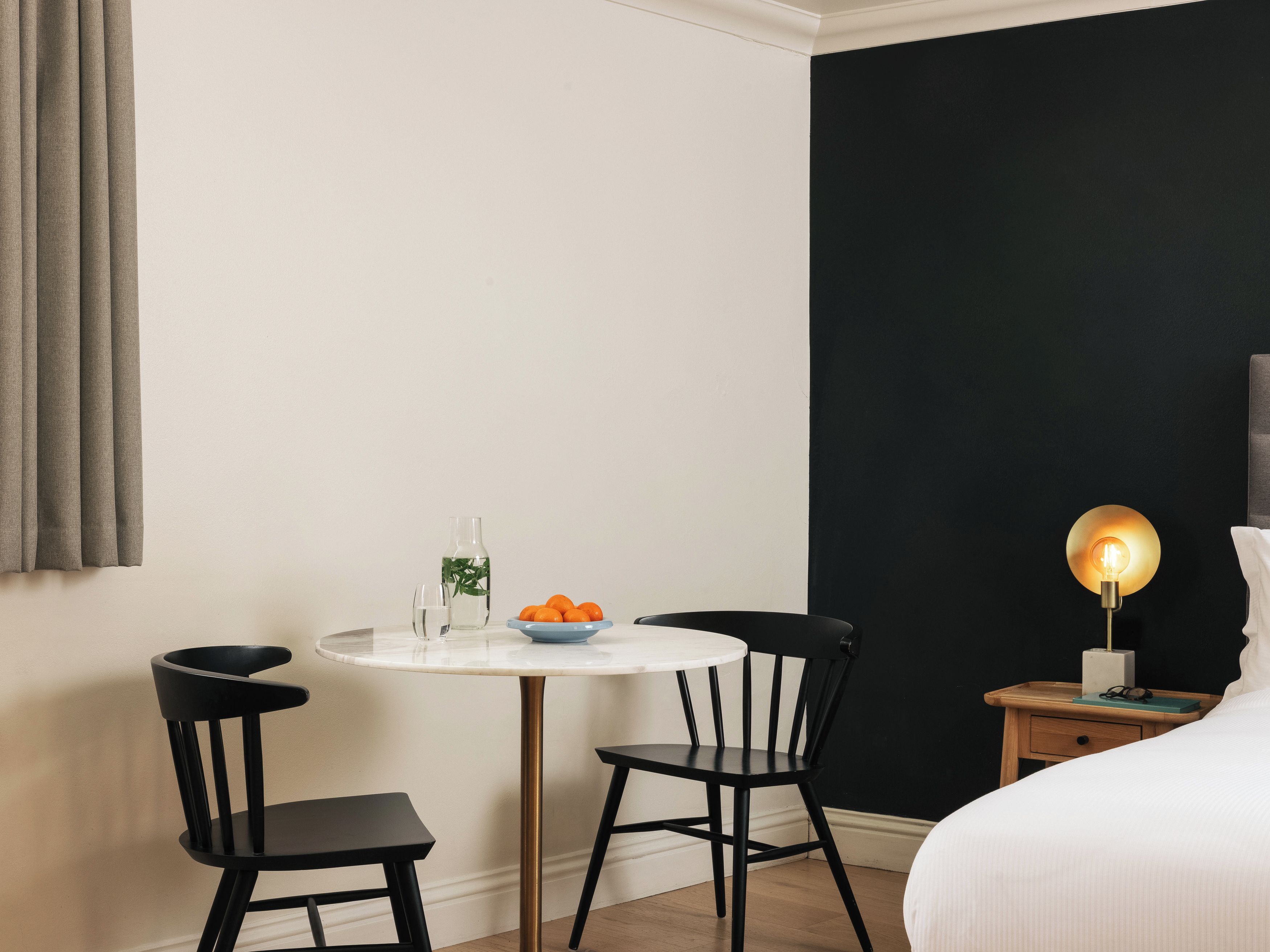 A hotel room with a round table, two chairs, a bedside table with a lamp, and a bed.