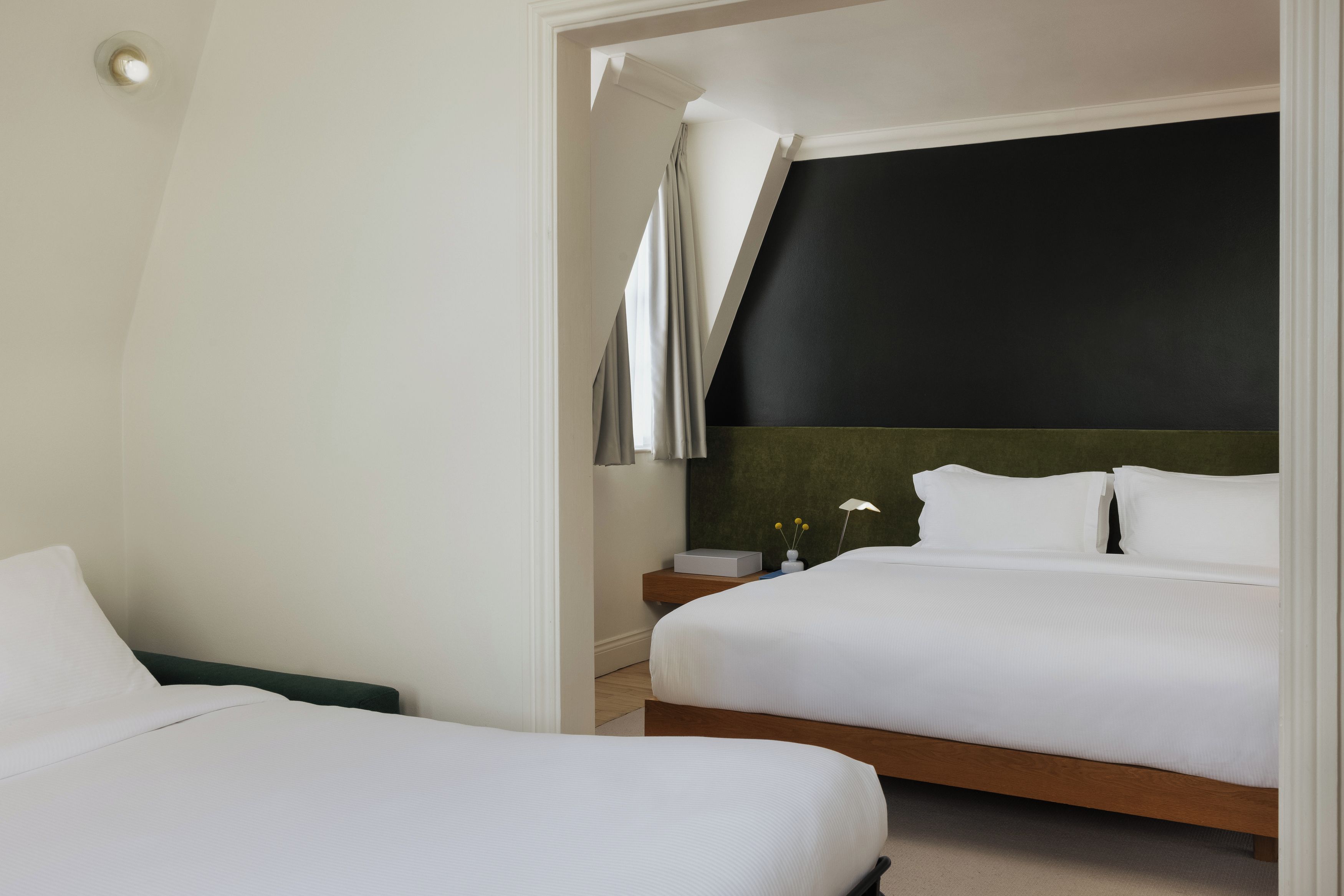A hotel room with two beds, a dark accent wall, a nightstand with a lamp, and a small table with a vase.