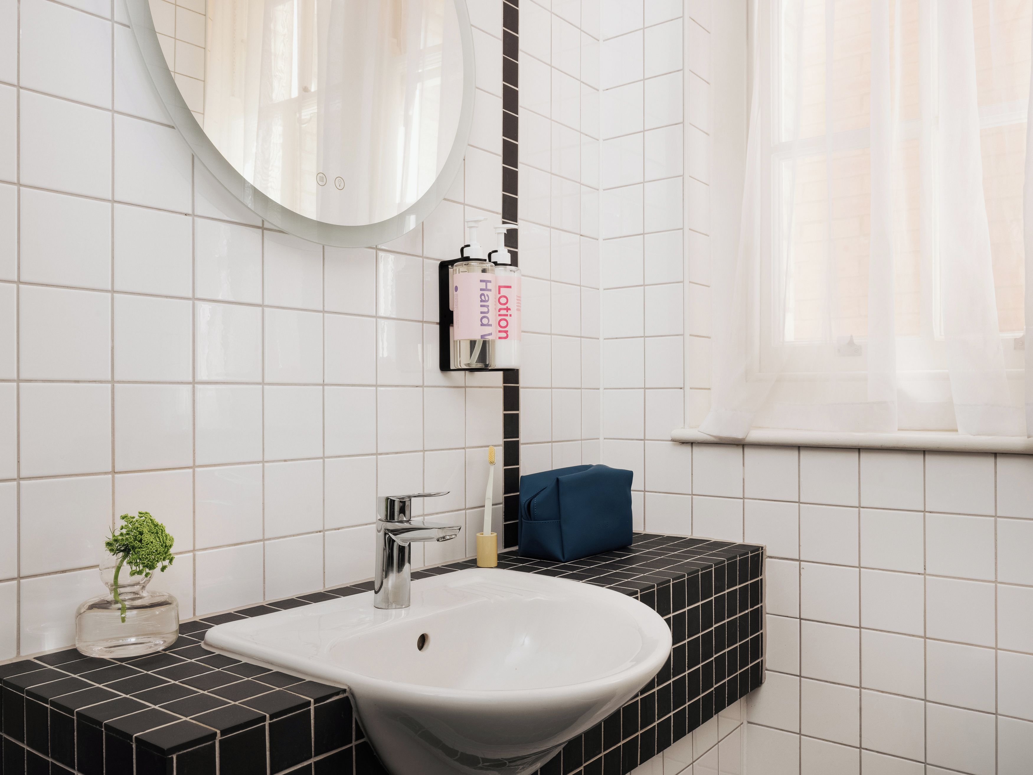 A bathroom with a white sink, black tiles, a mirror, and a window with sheer curtains.