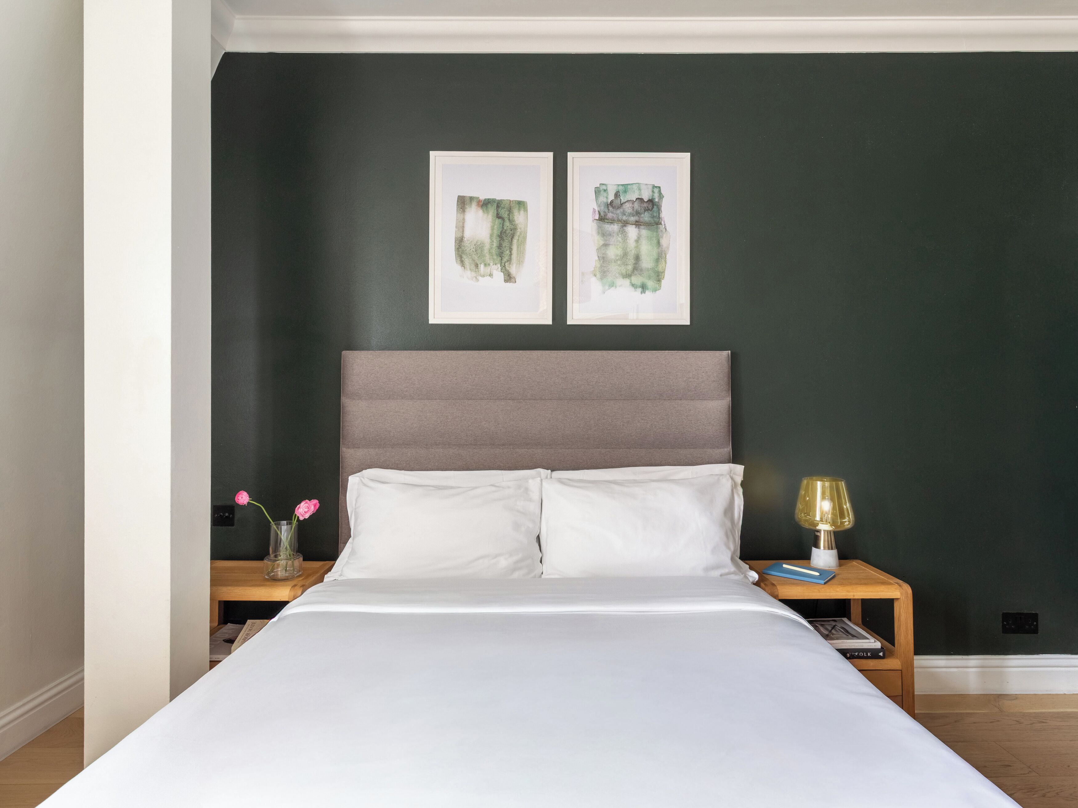 Quarto com cama, duas obras de arte emolduradas na parede, mesa de cabeceira com luminária e um vaso com flores.