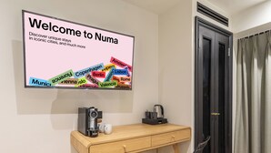 Una habitación de hotel moderna con un escritorio de madera, una silla negra, una cafetera y una pantalla mural que muestra un mensaje de bienvenida con destinos turísticos.