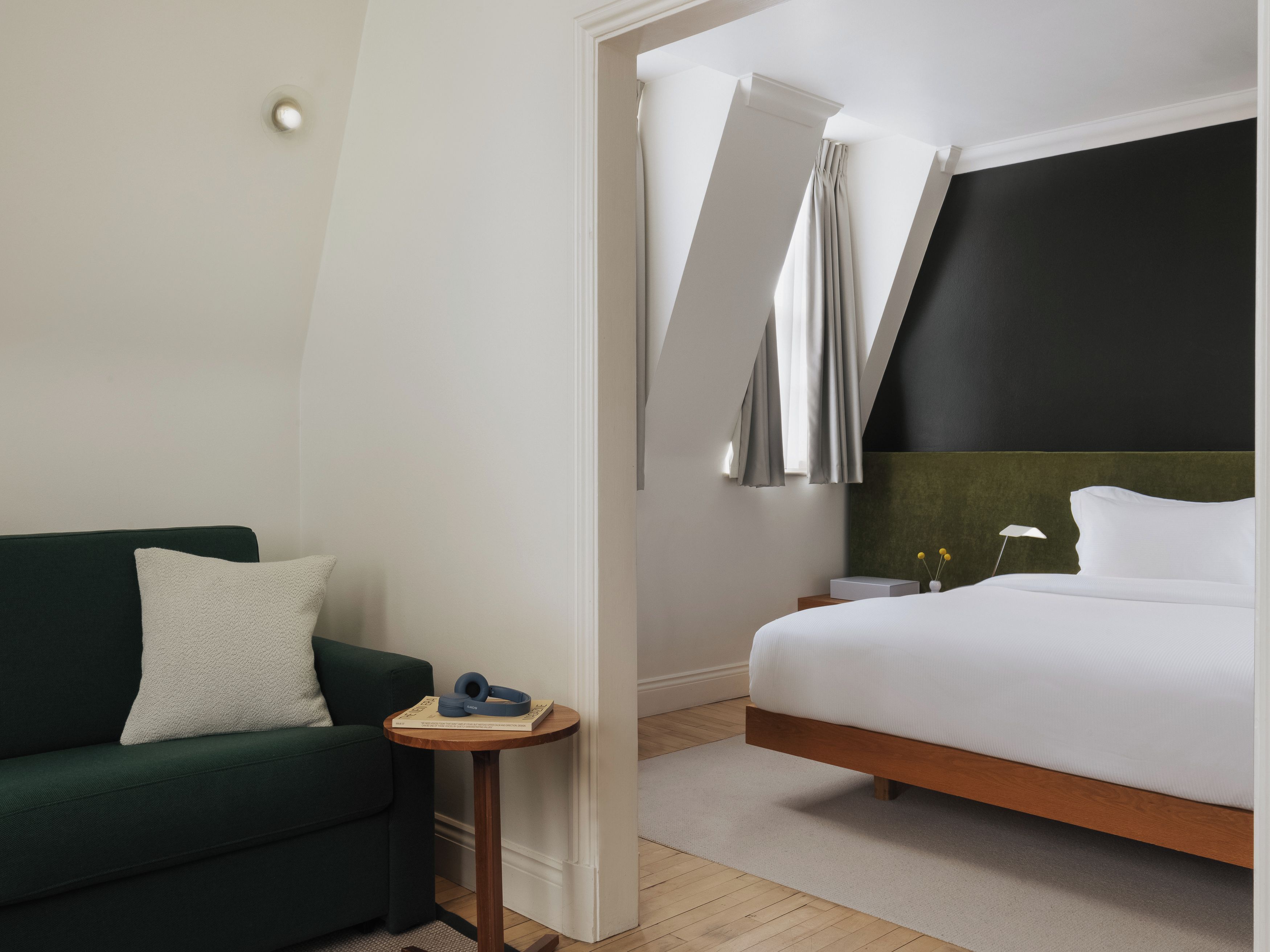 A modern hotel room with a green sofa, a wooden bedside table, and a bed with white bedding.