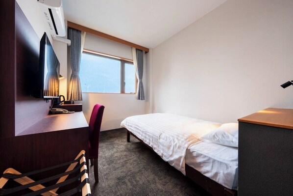 A hotel room with a bed, a desk with a lamp, a chair, and an air conditioning unit.