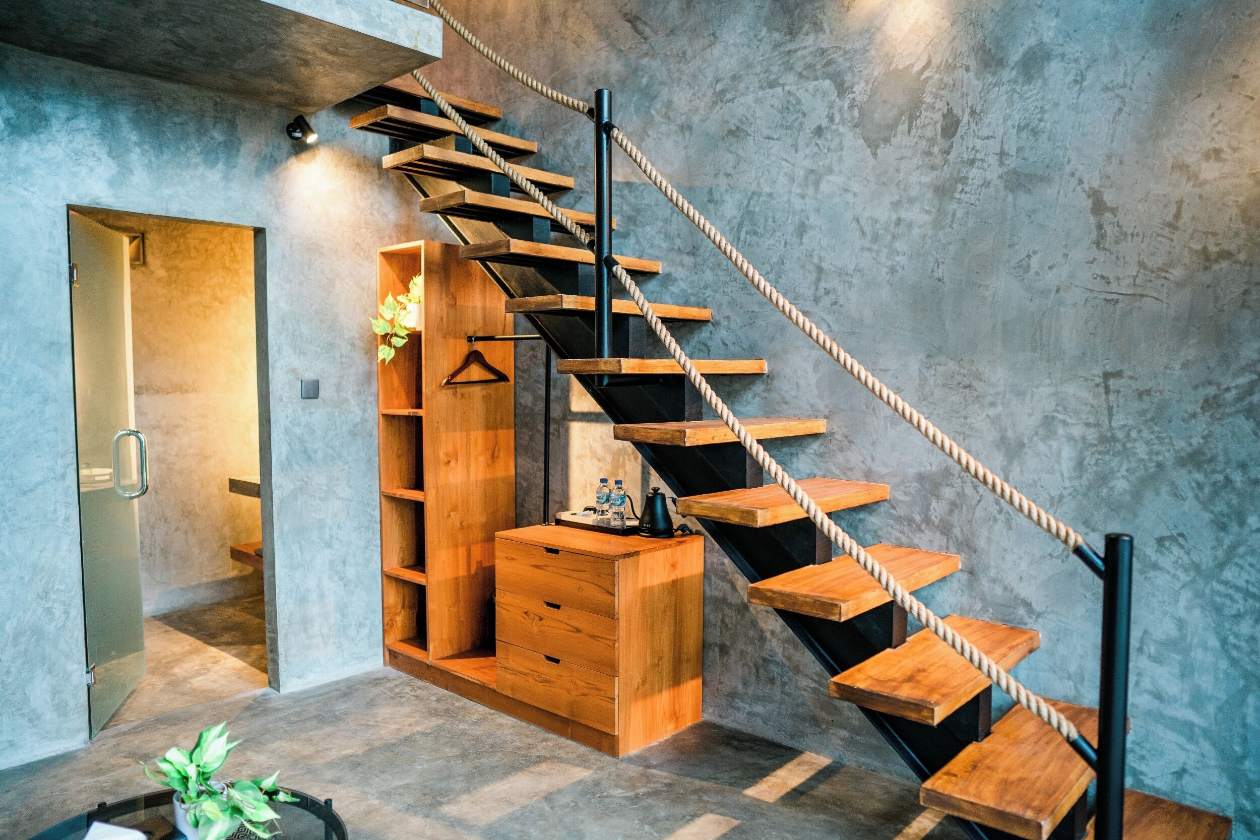 A modern staircase with wooden steps and a rope handrail, leading to a room with a wooden cabinet and a hanging plant.