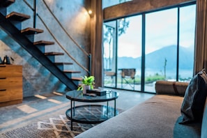 A modern living room with a sofa, a coffee table, a staircase, and a large window offering a view of the outdoors. - Maisaba Kintamani Cabin and Bistro (Kintamani)