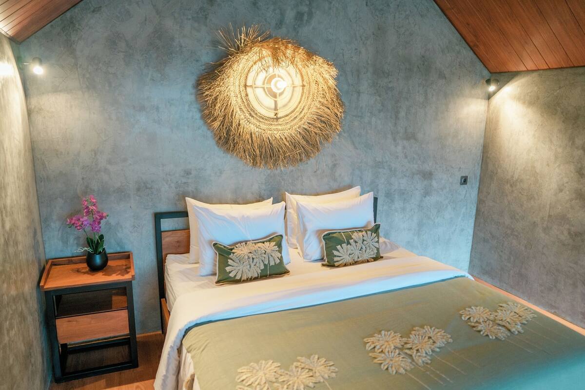 Una camera da letto con un letto, cuscini, un comodino, un elemento decorativo a parete e una piccola pianta.