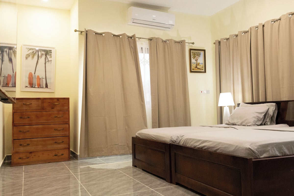 A bedroom with a wooden dresser, a bed with a headboard, beige curtains, and a wall-mounted air conditioner.