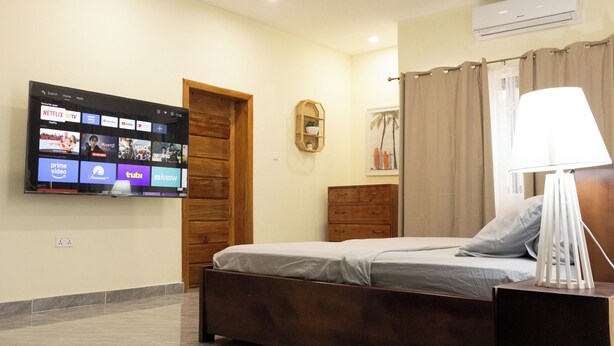Ein Schlafzimmer mit einem Bett, einem an der Wand montierten Fernseher, der Streaming-Dienste anzeigt, einem Holzschrank, einem Nachttisch mit Lampe und einem Fenster mit Vorhängen.