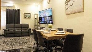 Ein Essbereich mit einem Holztisch, gedeckt für sechs Personen, umgeben von Stühlen, einer Couch und einem Fernseher, der einen Reisekanal zeigt.