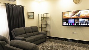 Ein Wohnzimmer mit grauem Ecksofa, einem gemusterten Teppich, einem an der Wand montierten Fernseher und einem schwarzen Regal mit einer Pflanze.
