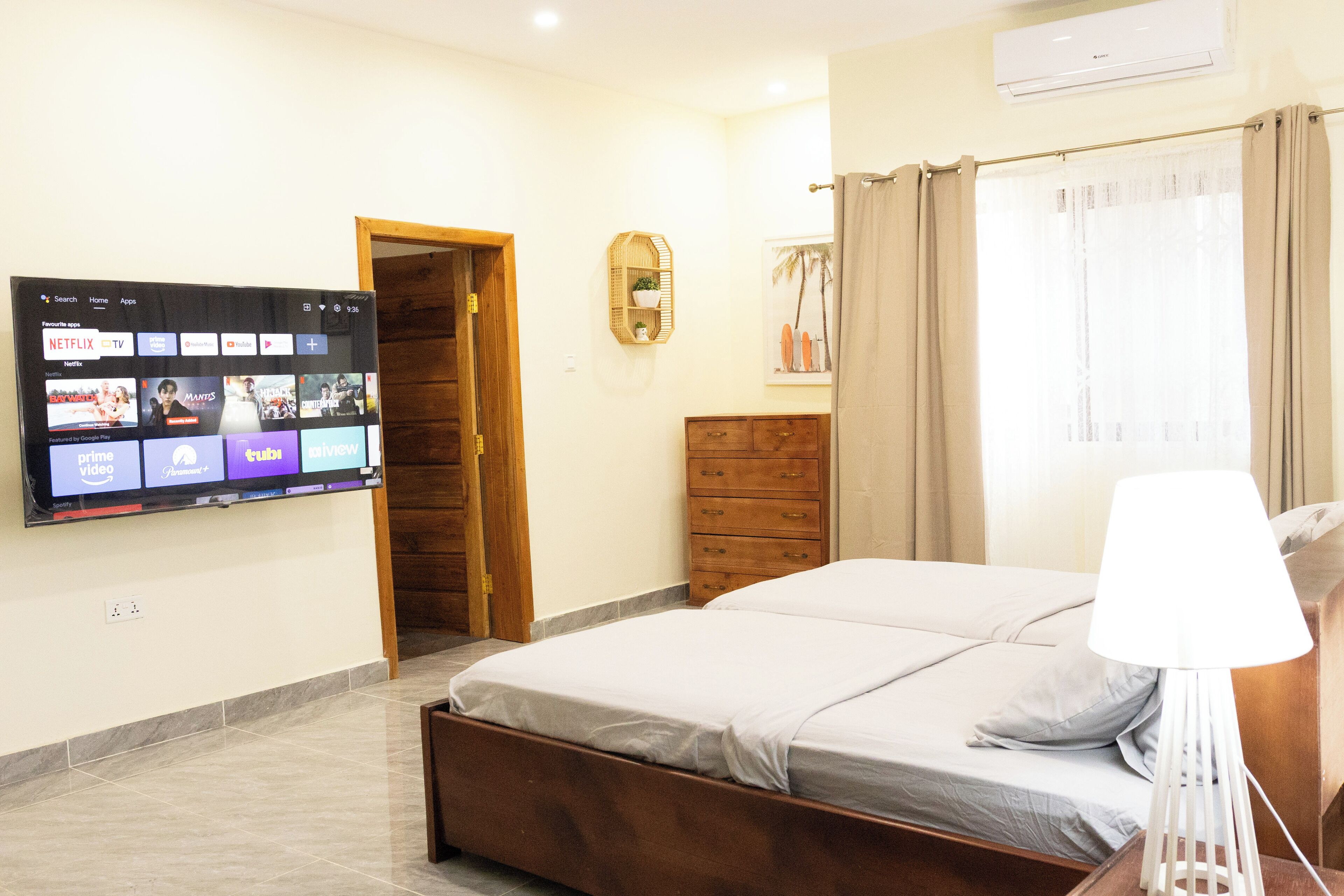 A hotel room with a bed, a TV mounted on the wall displaying various streaming services, a wooden door, a dresser, and a window with curtains.