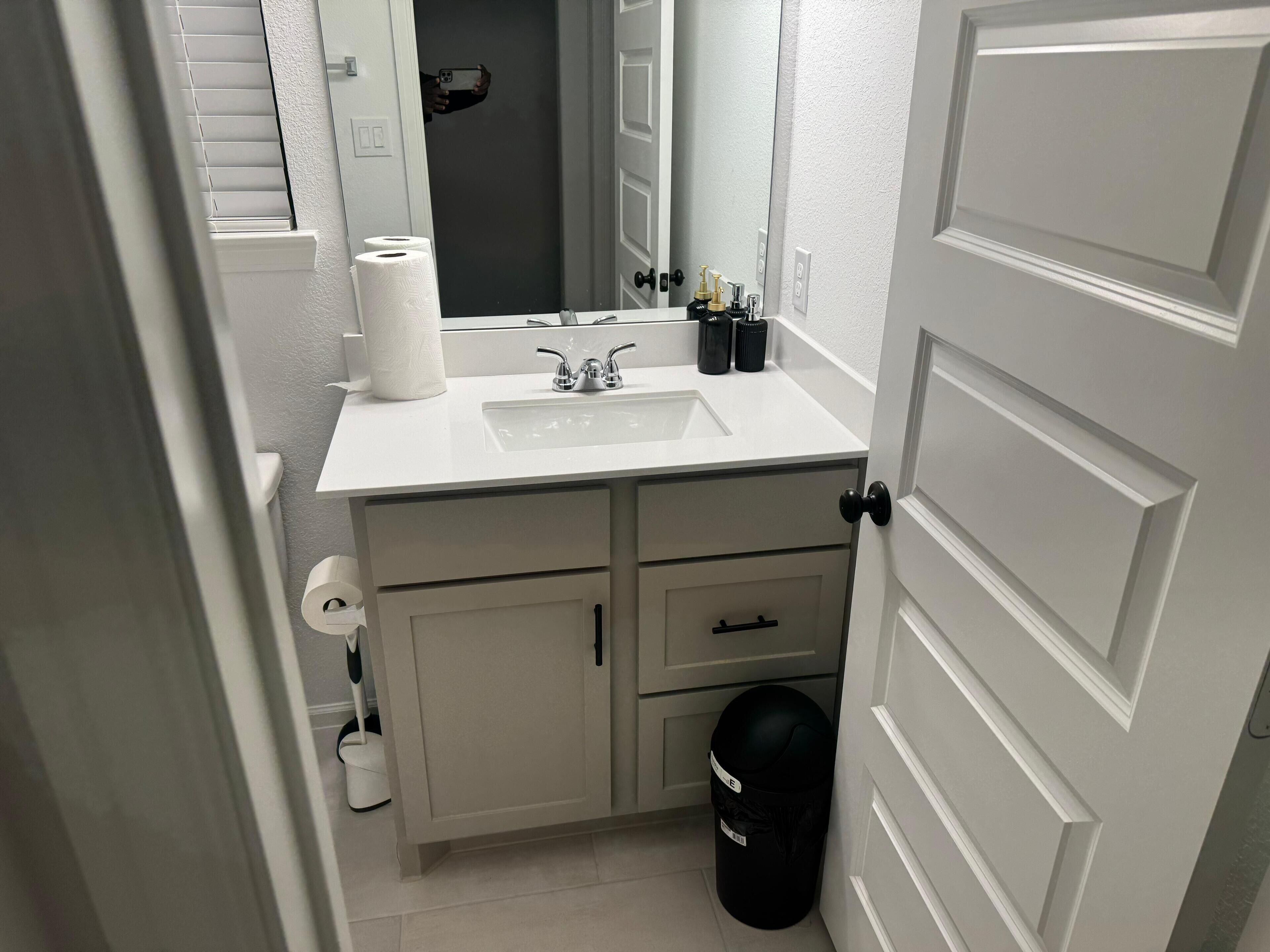 Een badkamer met een wit wastafel, spiegel en twee zeepdispensers. Er is een zwarte prullenbak en een toiletpapierhouder aan de muur.