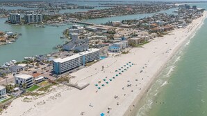 Ein Luftbild einer Küstenstadt mit einem langen Sandstrand, einem Jachthafen und mehreren Gebäuden, darunter ein Hotel mit Pool und Strandstühlen.