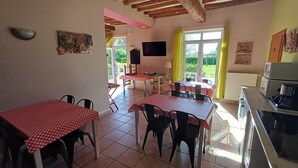 Un coin repas avec des tables à carreaux rouges et blancs, une cuisine équipée d’une cuisinière et une vue sur la verdure par la fenêtre.