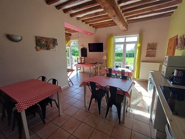 A dining area with red and white checkered tables, a kitchen with a stove, and a view of greenery through the window. - Le Clos Saint Vincent & SPA (Manneville-ès-Plains)
