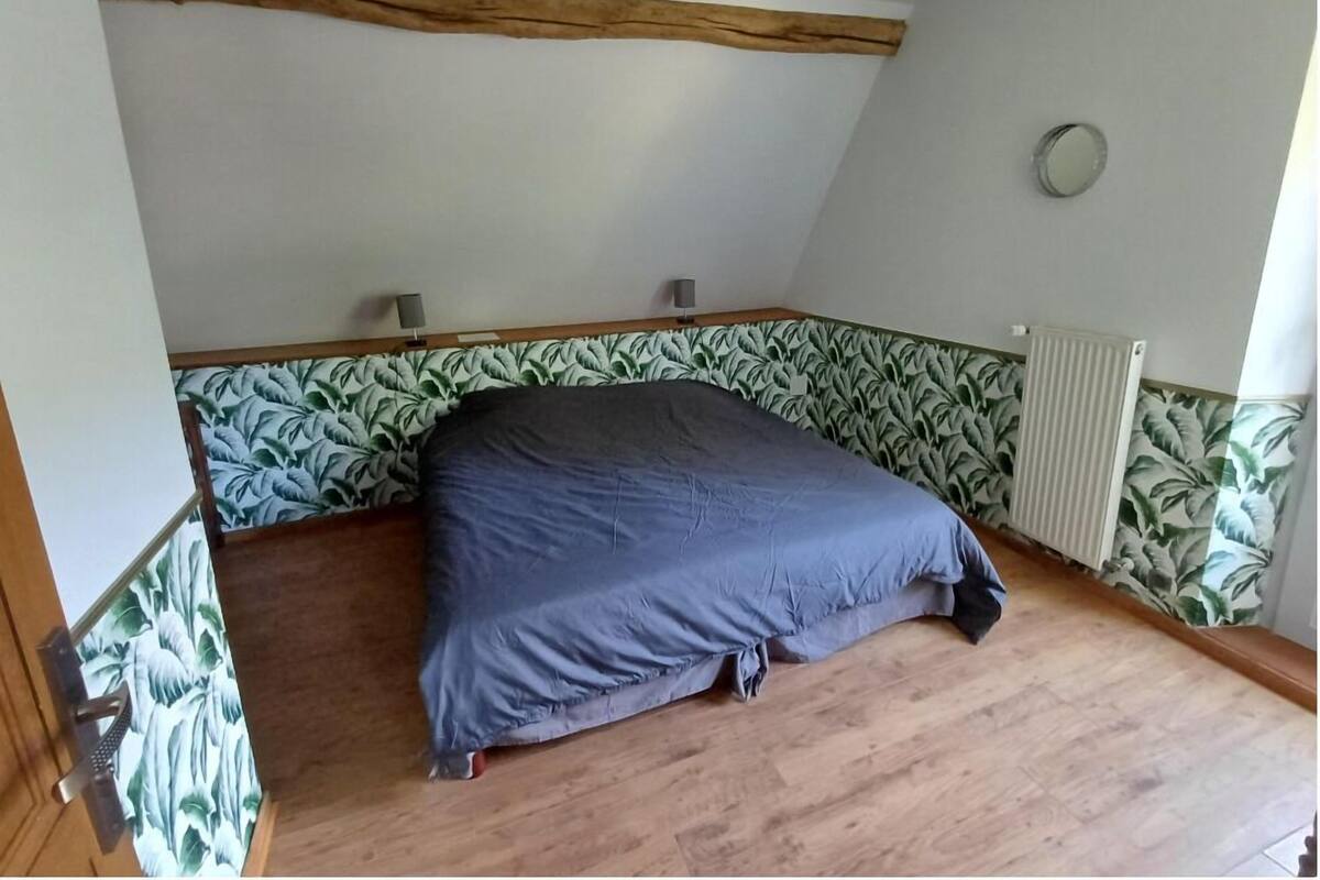 Une chambre avec un plafond en pente, un lit recouvert d’une couverture grise, un radiateur et un papier peint à motifs sur les murs.