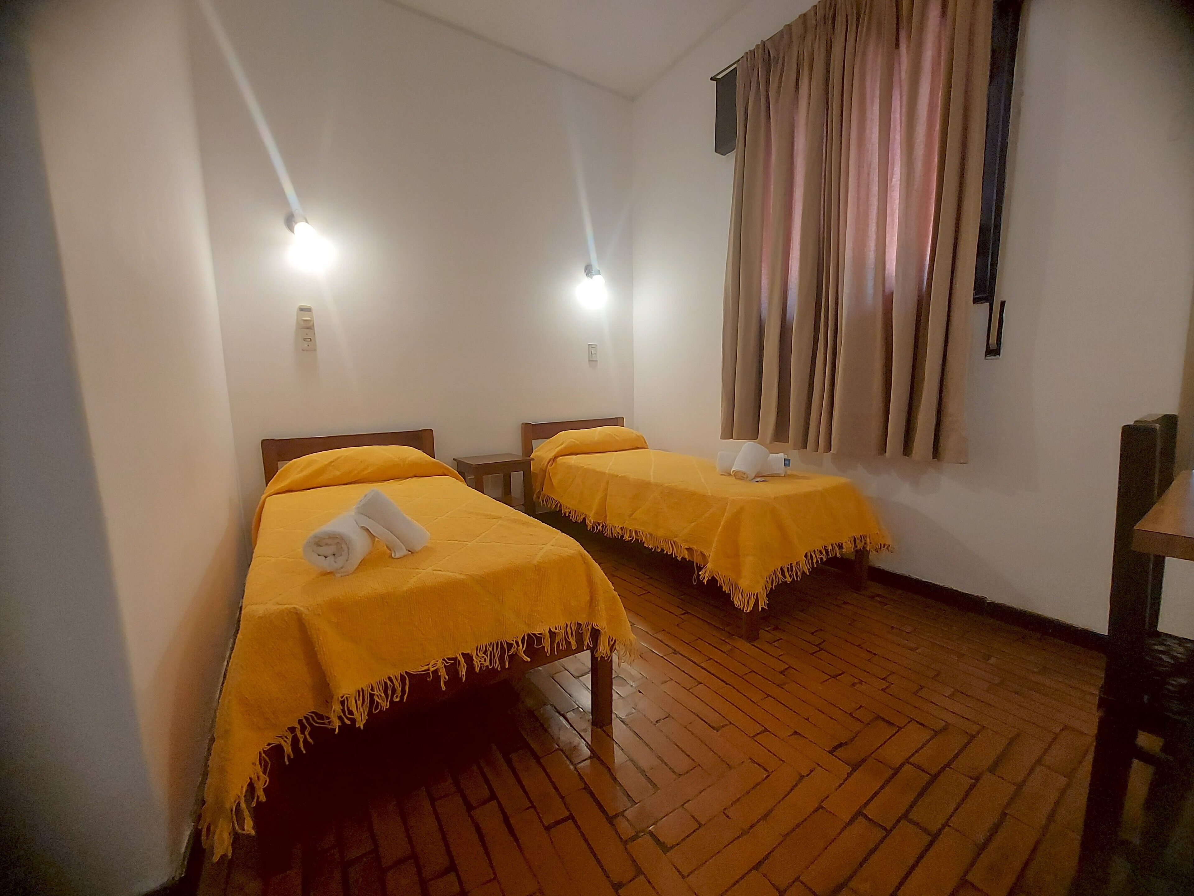 A room with two beds, each with a yellow bedspread and a rolled towel on the bed. There is a wooden floor and a window with curtains.