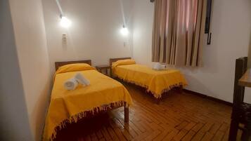 A room with two beds, each with a yellow bedspread and a rolled towel on the bed. There is a wooden floor and a window with curtains.