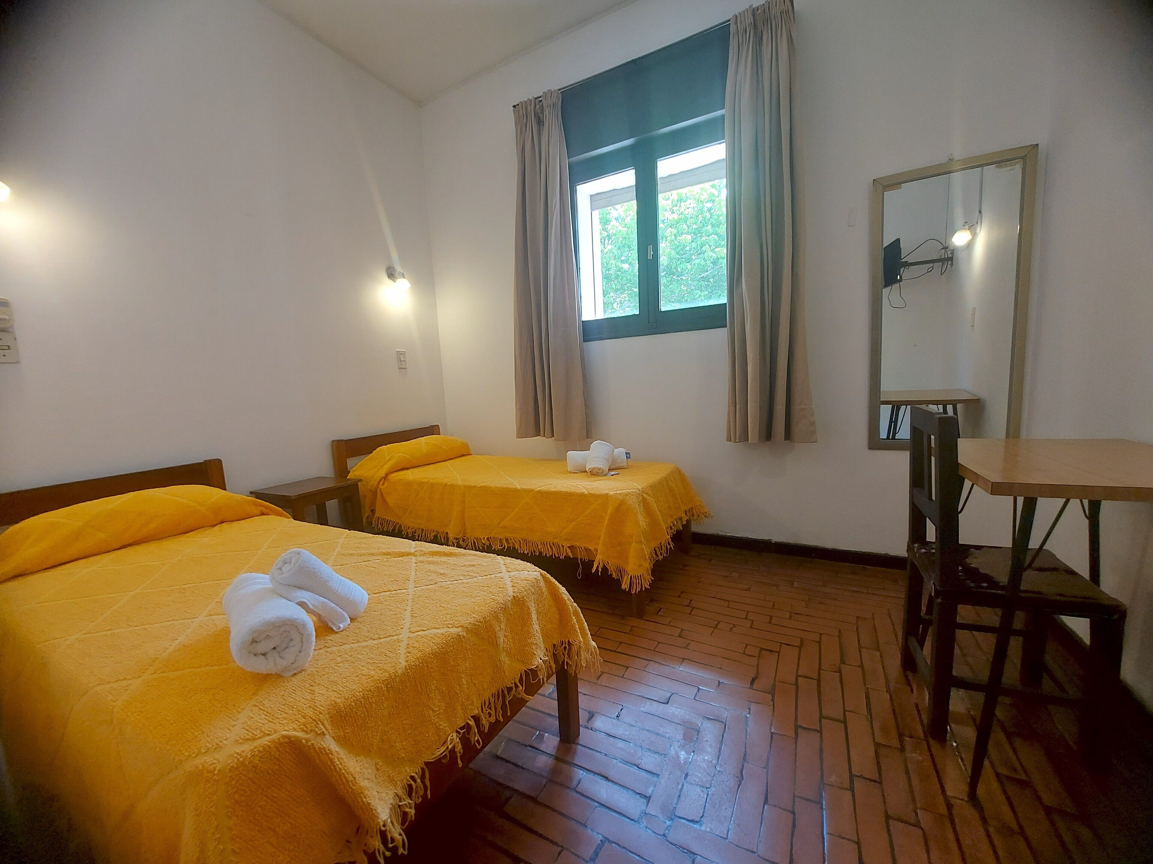Une chambre d’hôtel avec deux lits, une table en bois, un miroir et une fenêtre avec des rideaux.