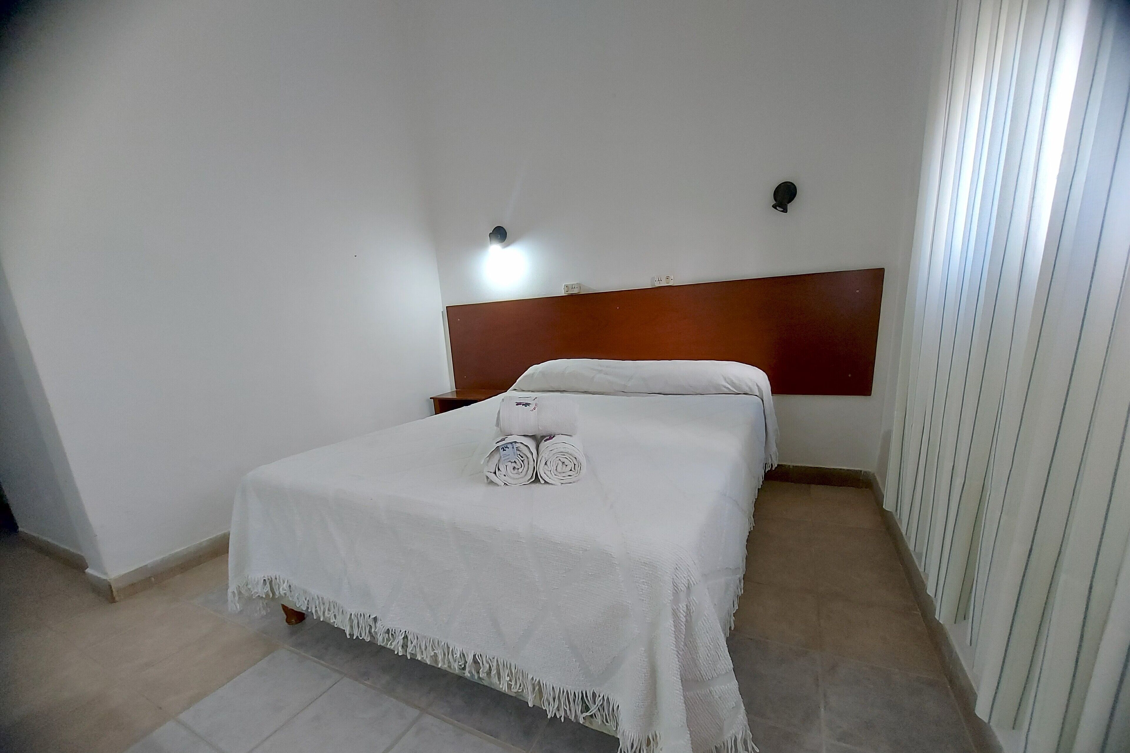 Habitación de hotel con una cama individual, cabecera de madera, colcha blanca y una mesita de noche con lámpara.