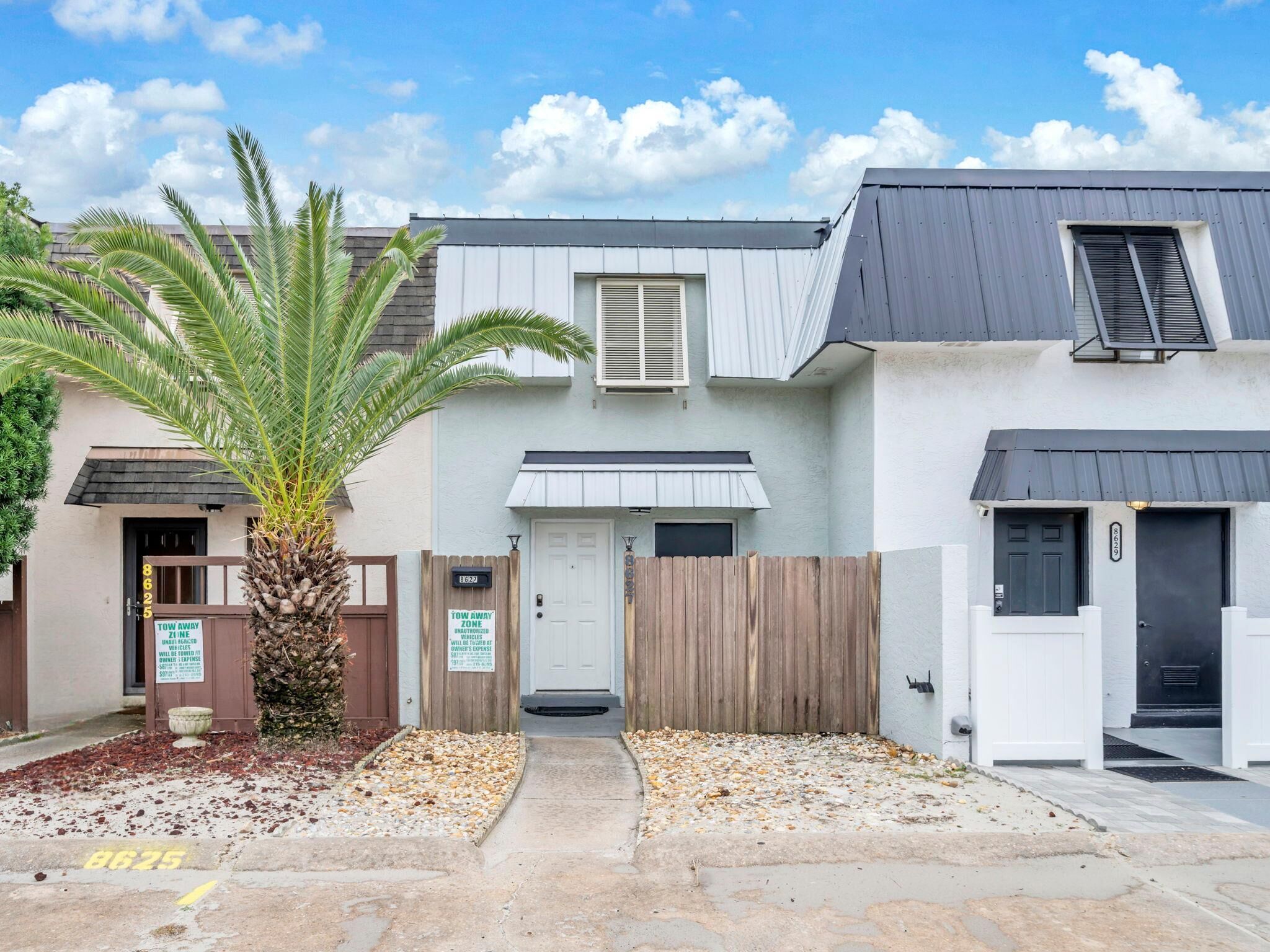A two-story residential building with a gated driveway, a palm tree, and a 'Tow Away Zone' sign.