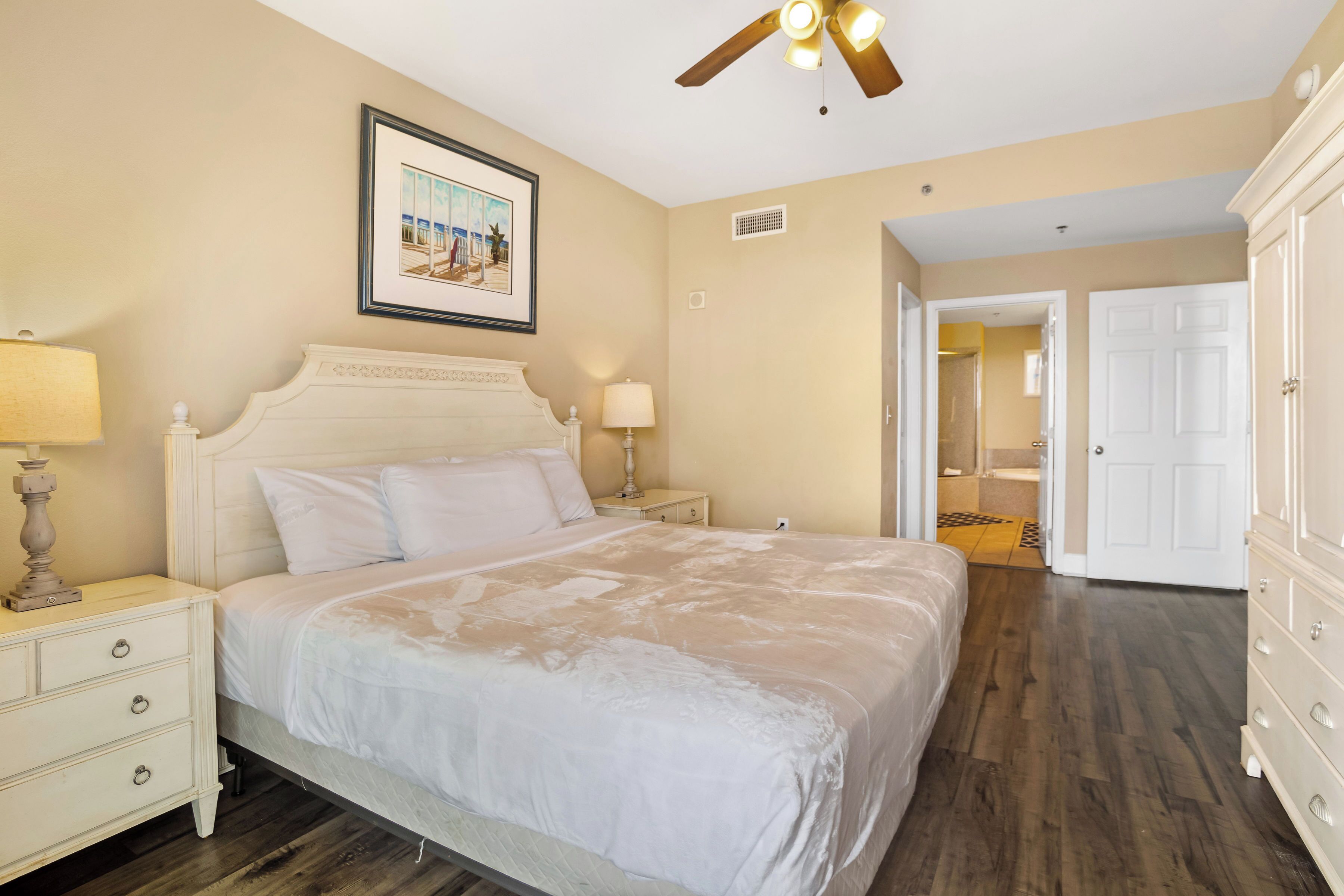 A bedroom with a large bed, two bedside tables, a ceiling fan, and a door leading to a bathroom.