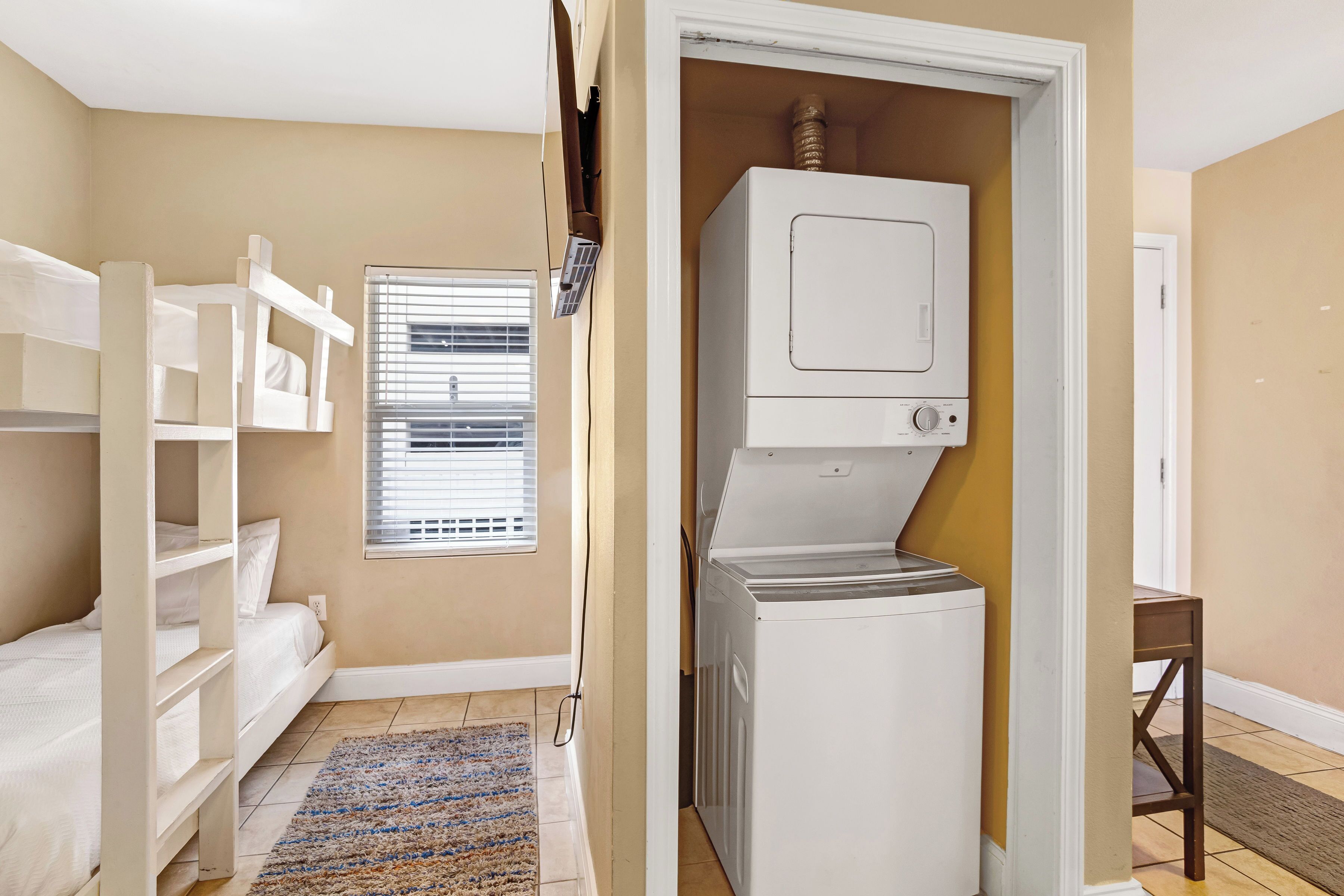 Une buanderie avec un lave-linge et un sèche-linge blancs, une fenêtre avec des stores, un lit superposé avec une literie blanche et un petit tabouret en bois.