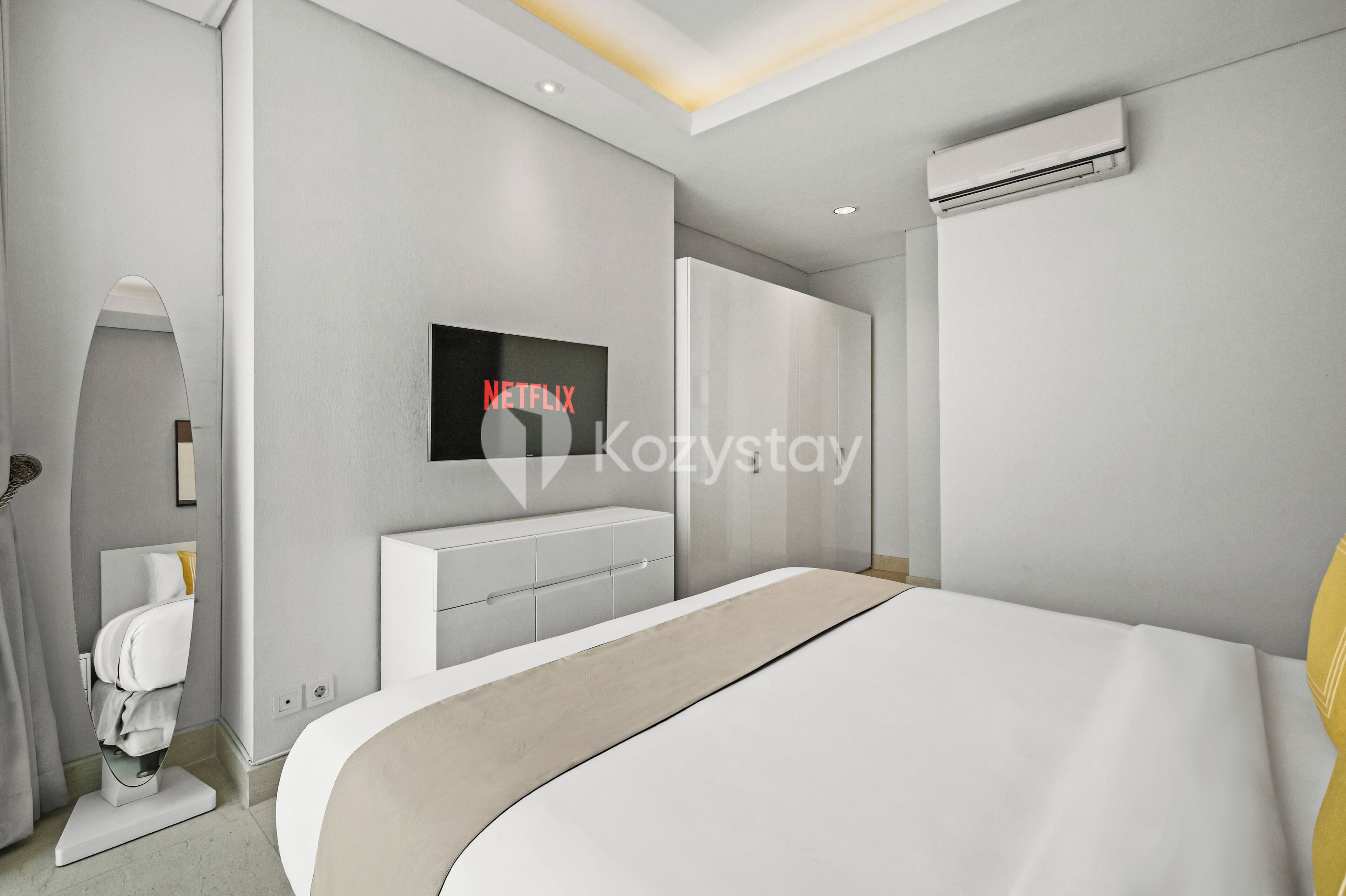 A modern bedroom with a large bed, a built-in TV, a white wardrobe, and an air conditioning unit.