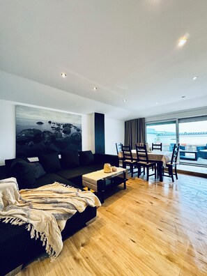 Sala de estar moderna com sofá preto, mesa de centro e área de jantar com cadeiras e mesa. Há uma pintura grande na parede e vista para o exterior pelas janelas.