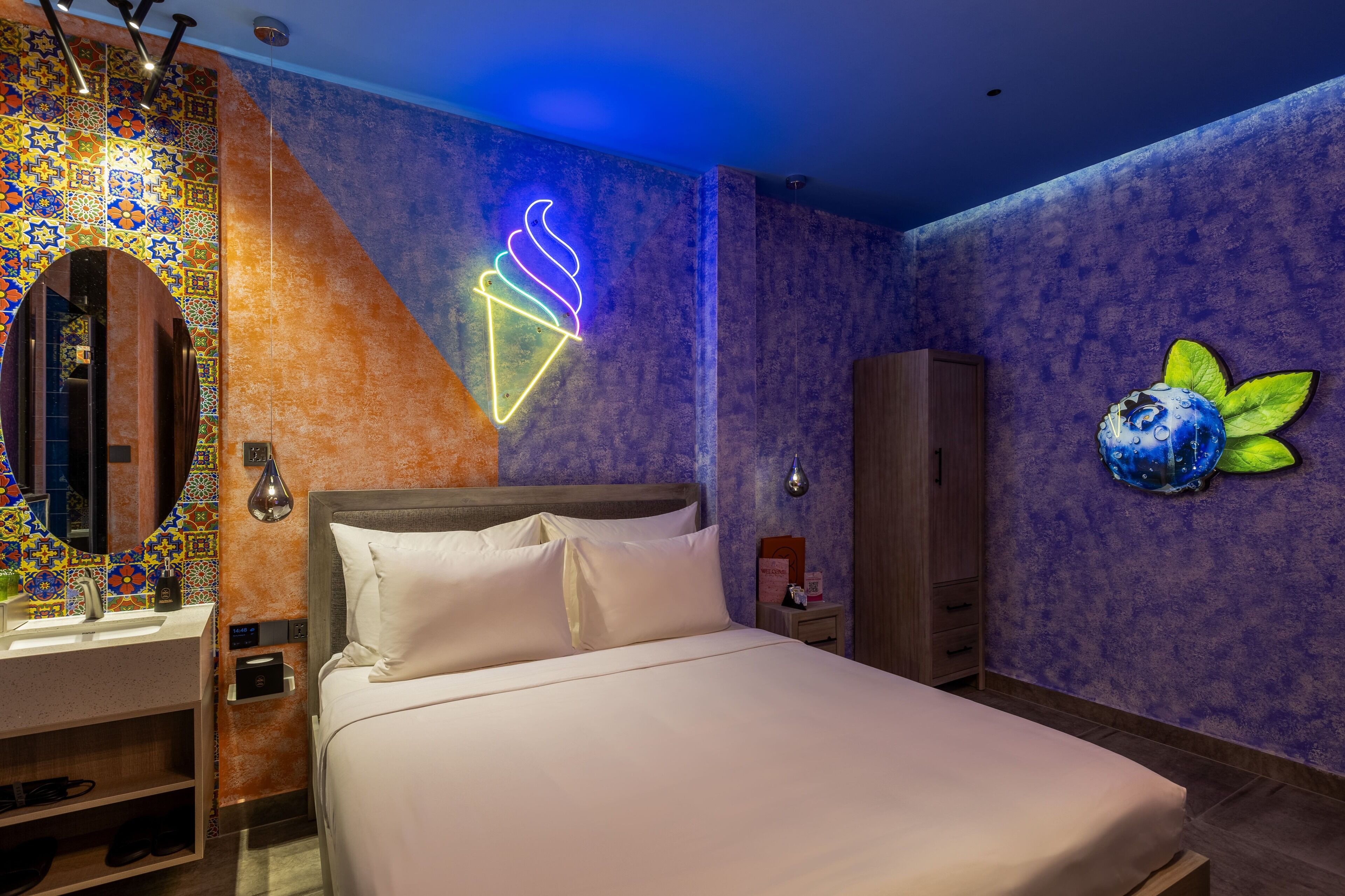 Une chambre d'hôtel avec un lit, un panneau lumineux en forme de cornet de glace, un mur décoratif et une œuvre d'art représentant des fruits.