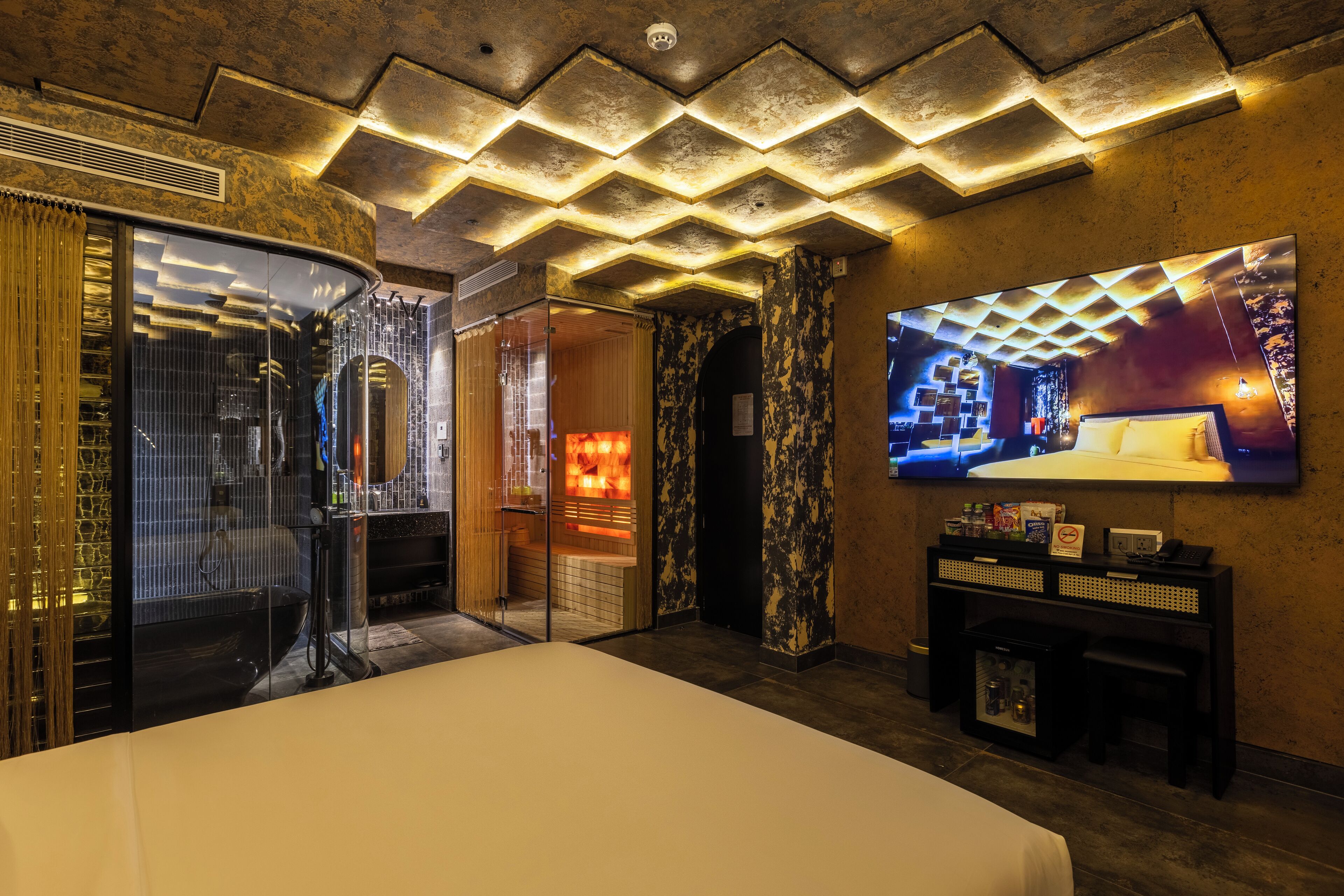 A hotel room with a large bed, a television mounted on the wall, a minibar, and a bathroom with a glass door.