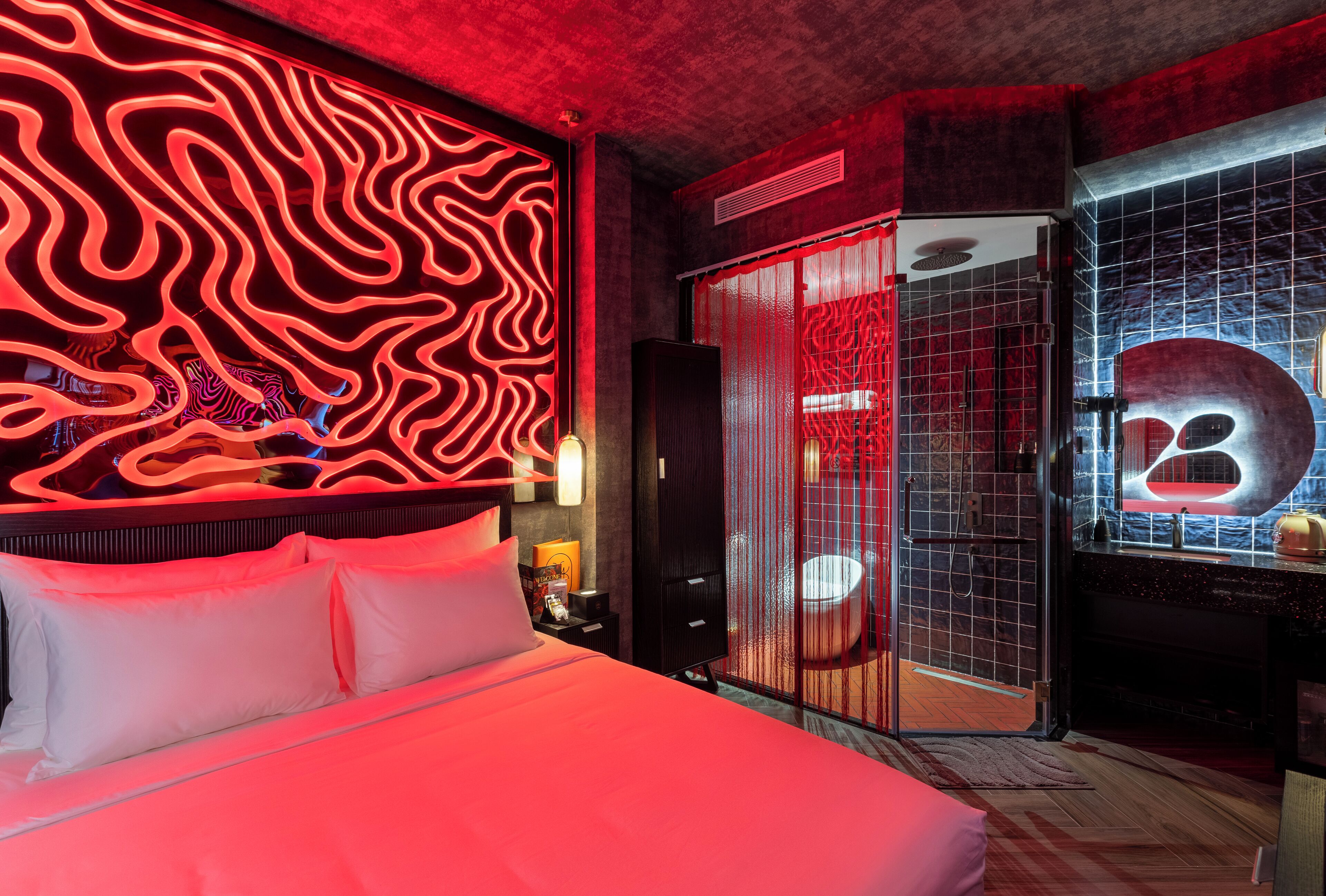 A modern hotel room with a red neon wall design, a large bed, a bathroom with a unique sink design, and a red and black color scheme.