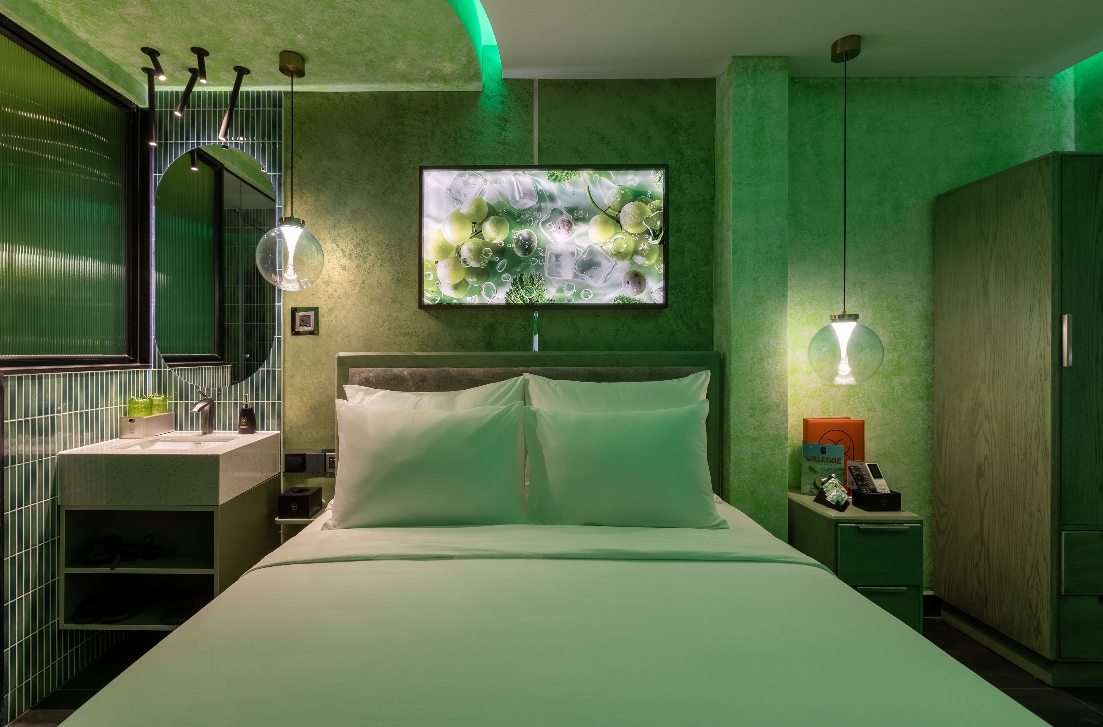 Una habitación de hotel moderna con decoración en tonos verdes, una cama grande, una mesita de noche con lámpara, un televisor y un armario.