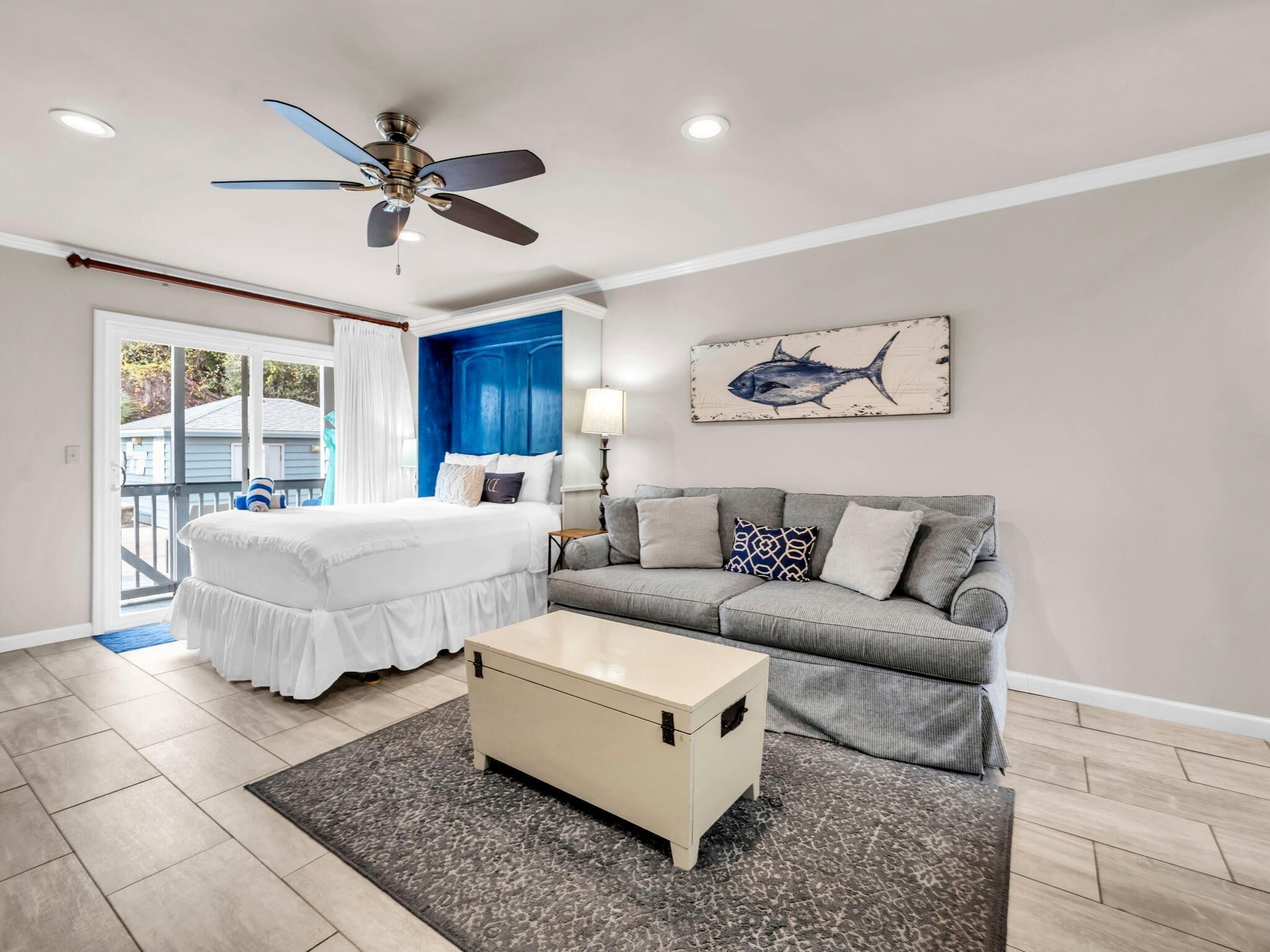 A bedroom with a bed, a gray sofa, a small table, a ceiling fan, and a fish painting on the wall.