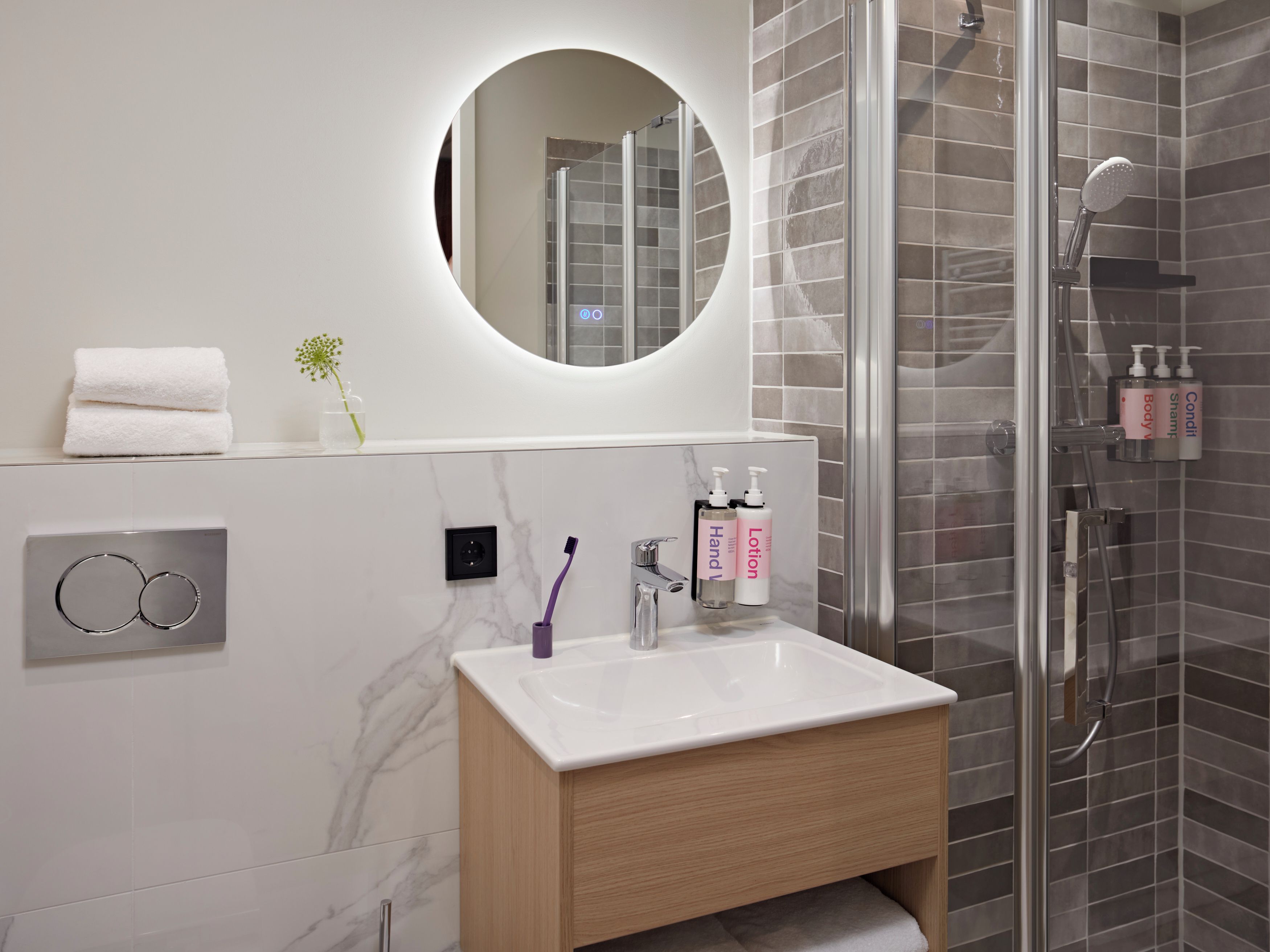 Une salle de bain moderne avec une douche à parois de verre, un mur recouvert de carreaux de marbre, un meuble-lavabo en bois et un miroir rond.