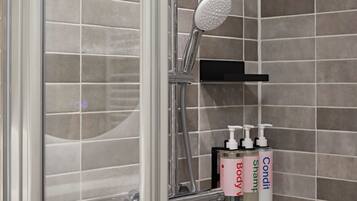 Une douche équipée d’un pommeau de douche à main, d’une porte en verre et d’une étagère où sont posés des flacons de gel douche et de shampoing.