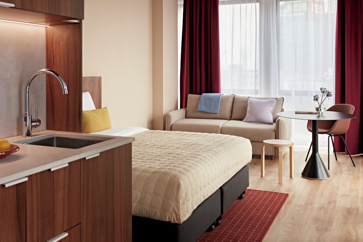 A modern hotel room with a kitchenette, a bed, a sofa, a small table, and a view of the city through large windows.