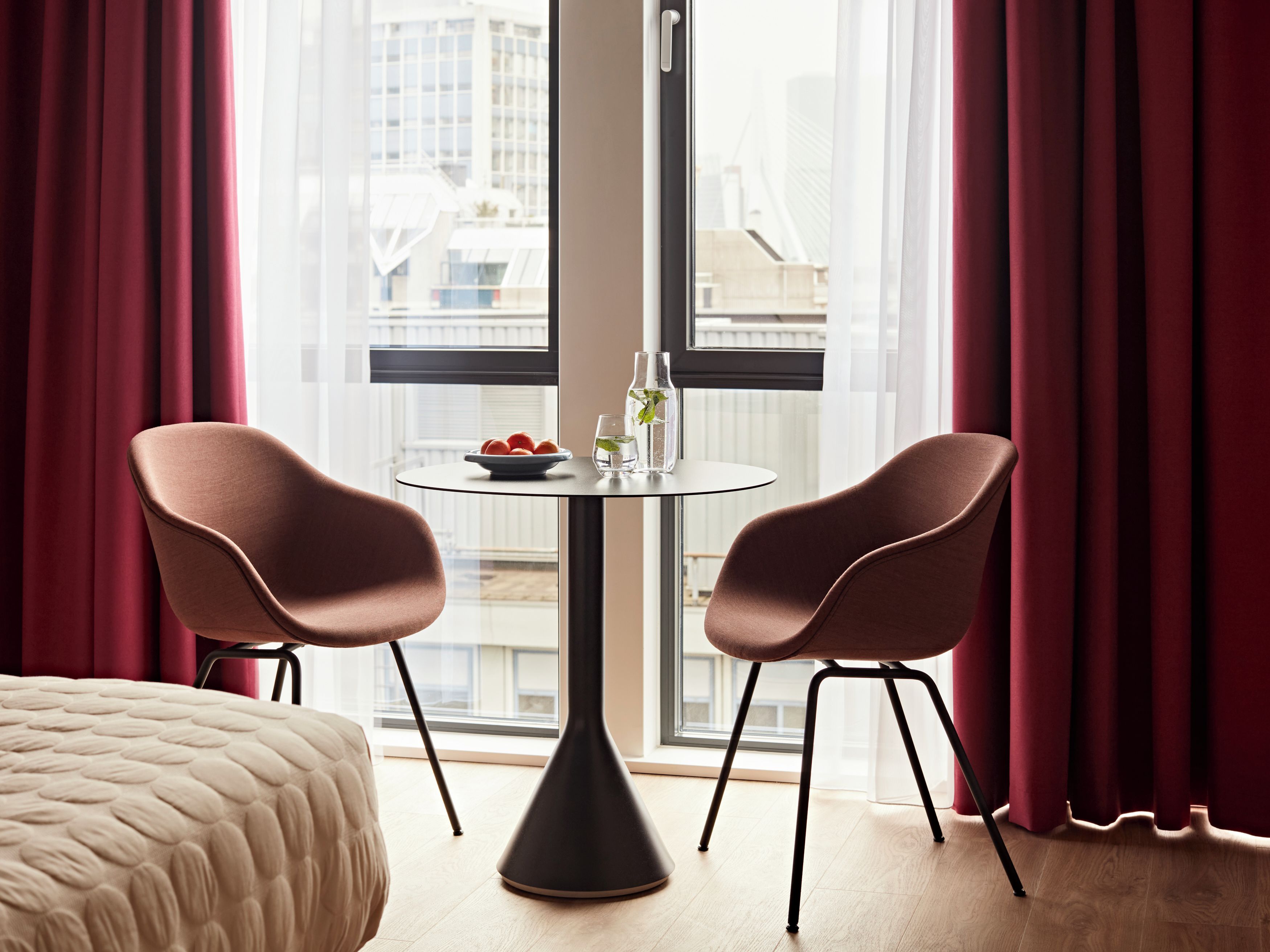 Una habitación de hotel moderna con un ventanal grande, dos sillas, una mesa redonda y una cama.