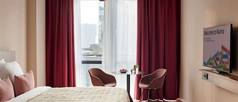 Une chambre d’hôtel avec un lit, deux chaises, une petite table avec une boisson, un téléviseur fixé au mur et une vue sur la ville à travers de grandes fenêtres.
