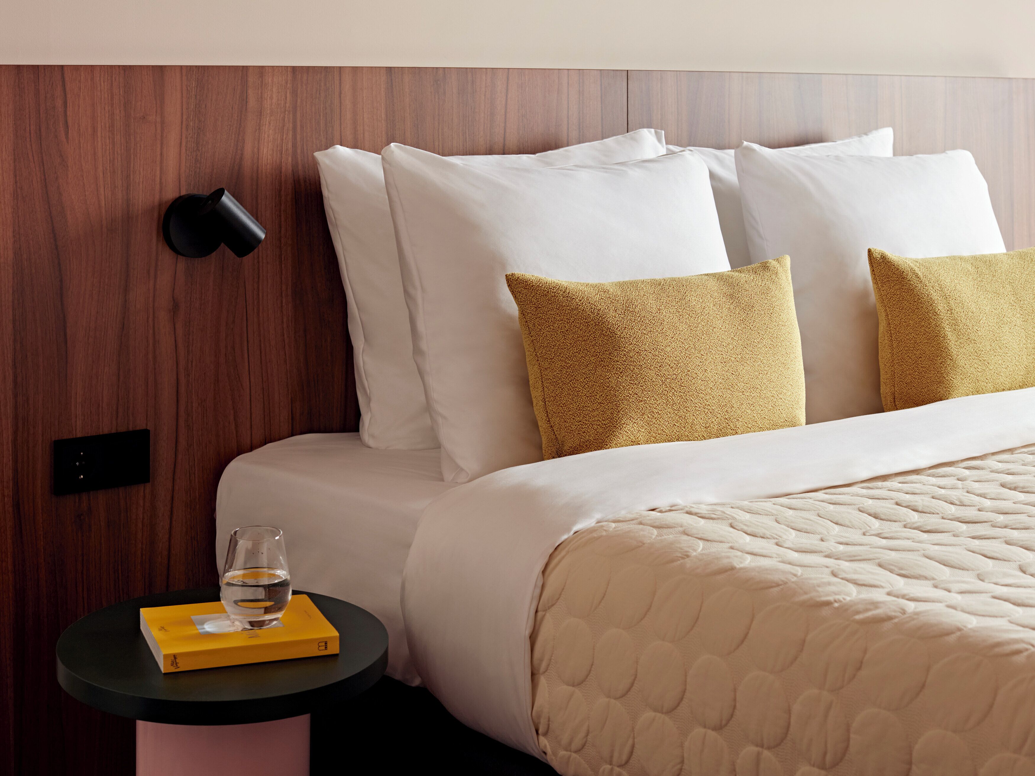 A neatly made bed with white and mustard pillows, a round bedside table with a glass of water and a book, and a wooden headboard.