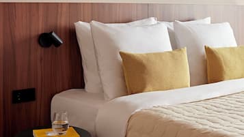 A neatly made bed with white and mustard pillows, a round bedside table with a glass of water and a book, and a wooden headboard.