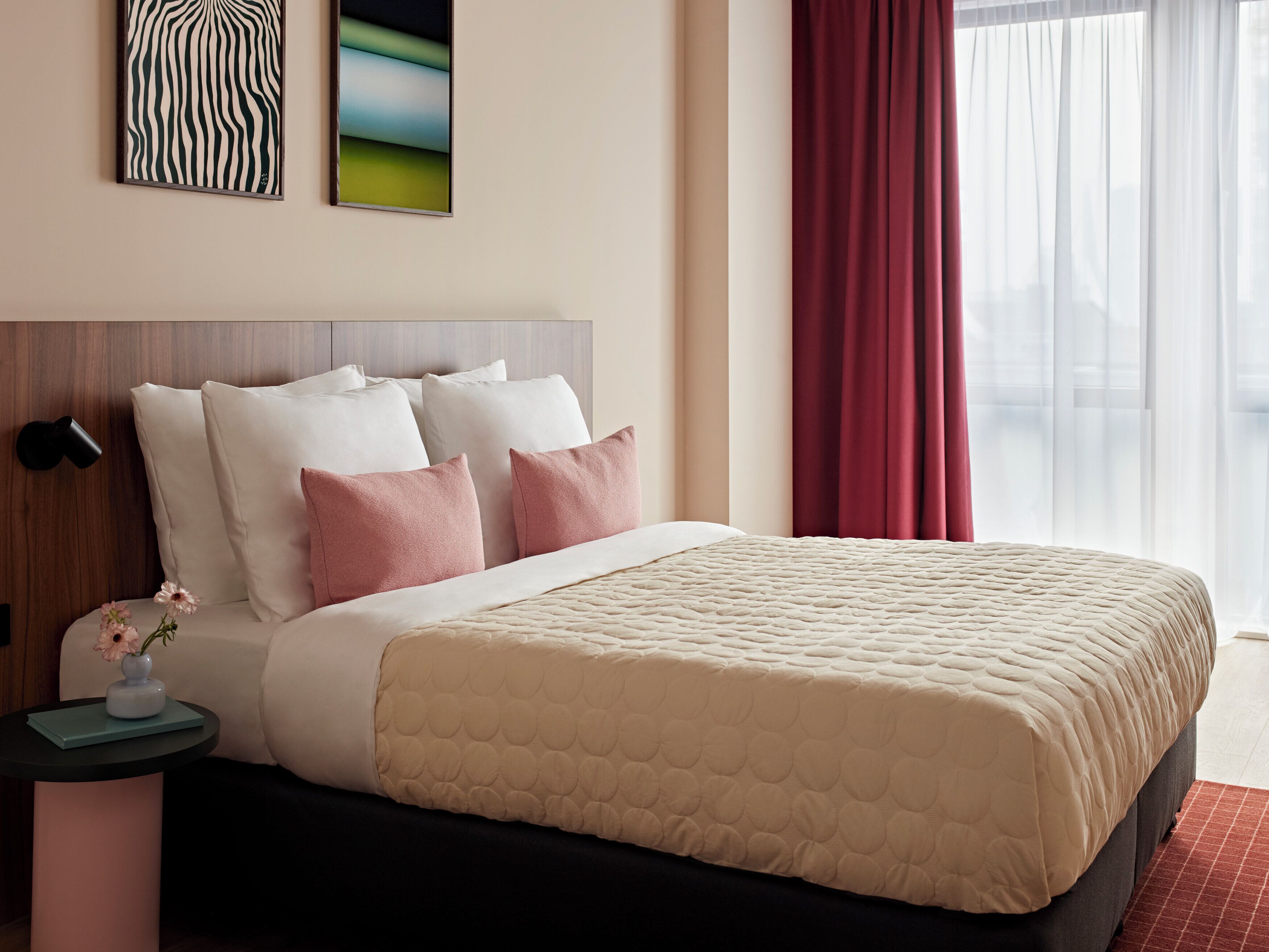 A hotel room with a bed, pillows, a nightstand, a vase with flowers, and artwork on the wall.