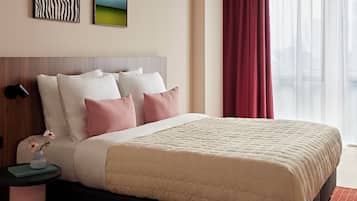 A hotel room with a bed, pillows, a nightstand, a vase with flowers, and artwork on the wall.