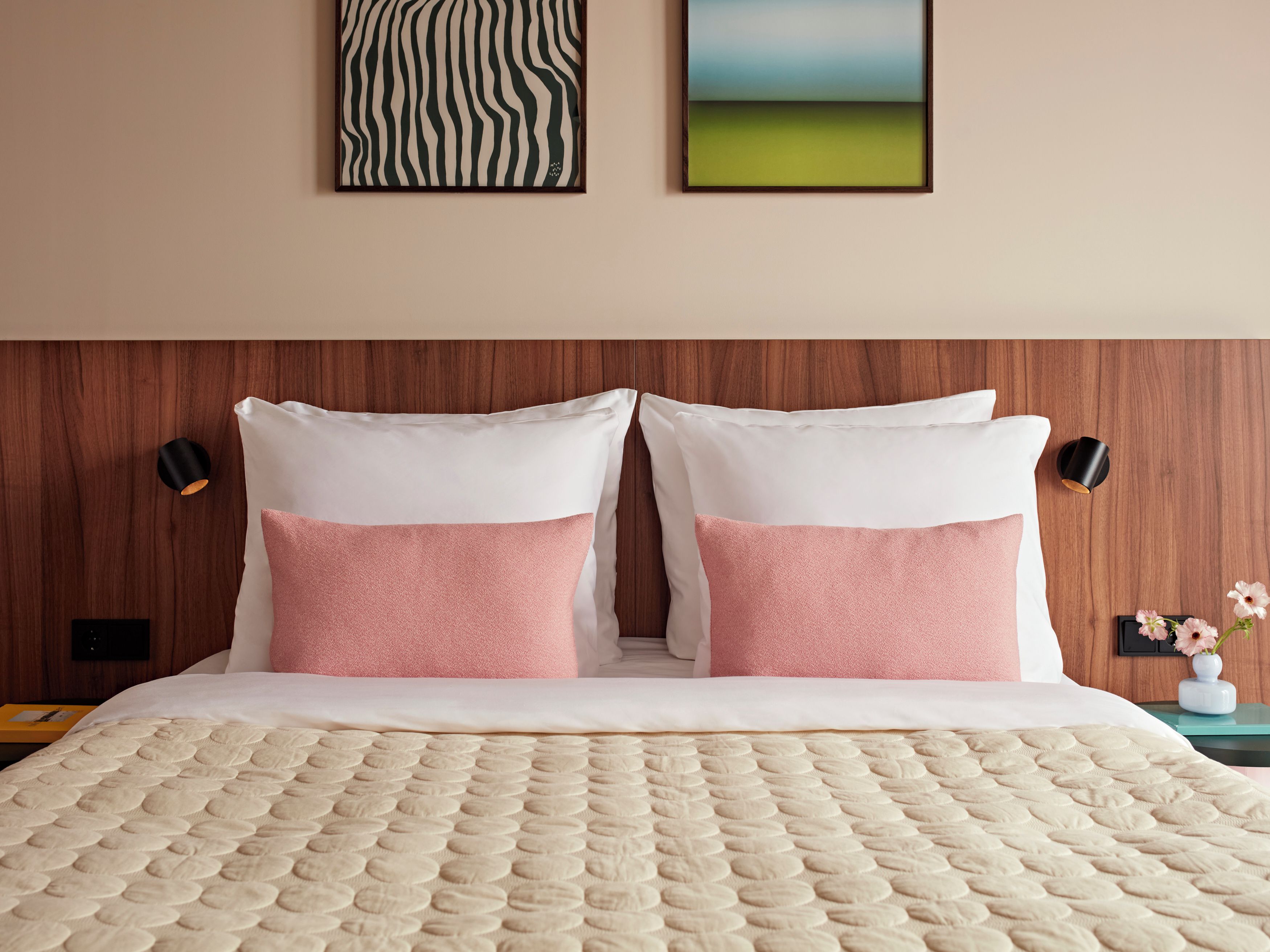 Une chambre d’hôtel avec une tête de lit en bois, deux oreillers blancs agrémentés de touches roses, une couvre-lit matelassée de couleur beige et deux lampes fixées au mur.